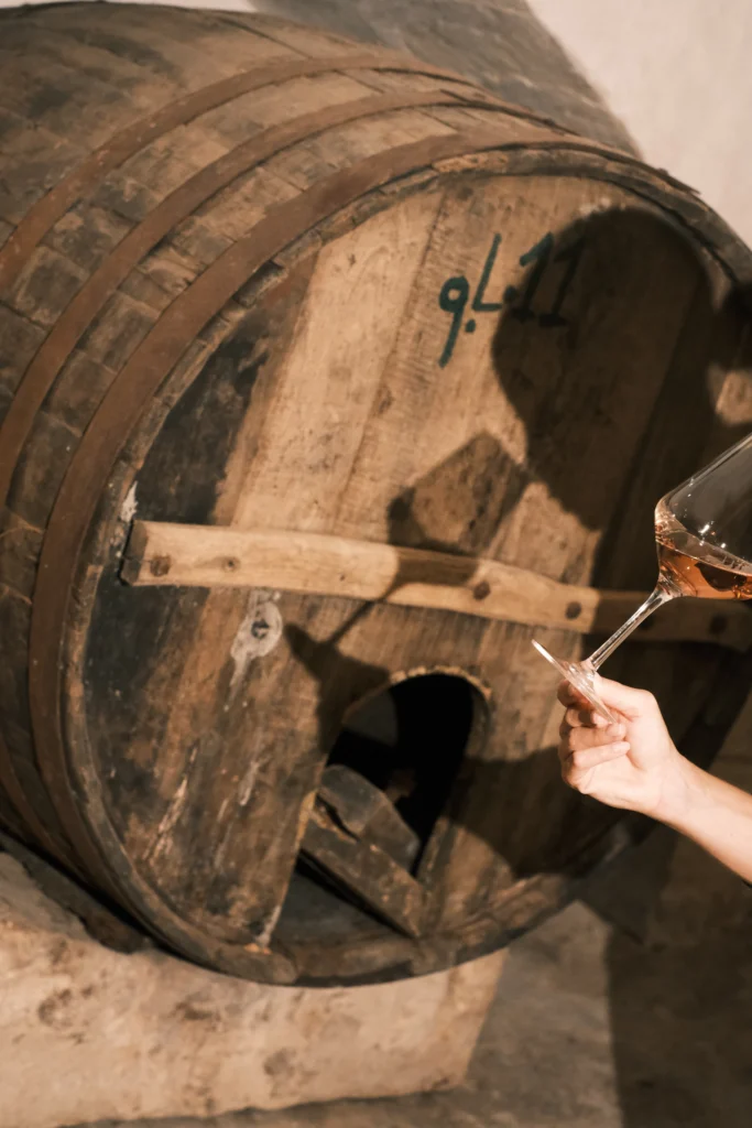 Glass of rosé wine held in front of a historic wooden barrel in a traditional Italian winery.