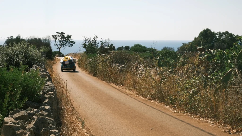 Vintage car driving on a narrow countryside road bordered by dry stone walls with distant sea view in Puglia