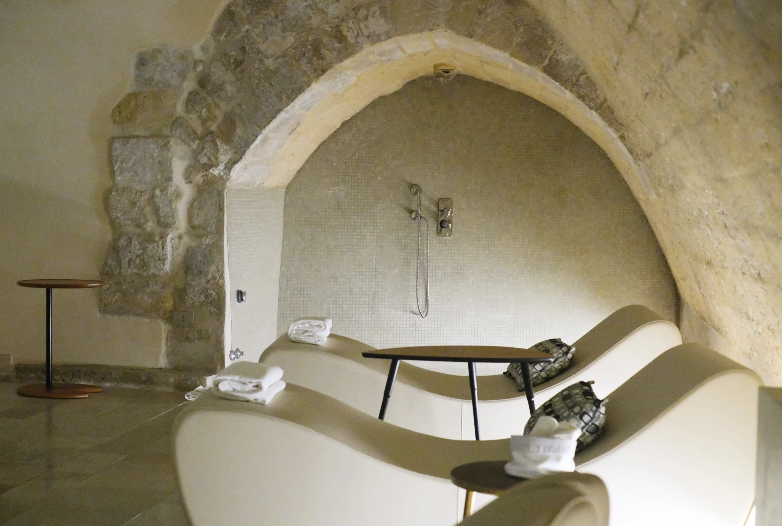 Stone-arched relaxation area with sculptural loungers and walk-in shower in a private underground spa in Puglia