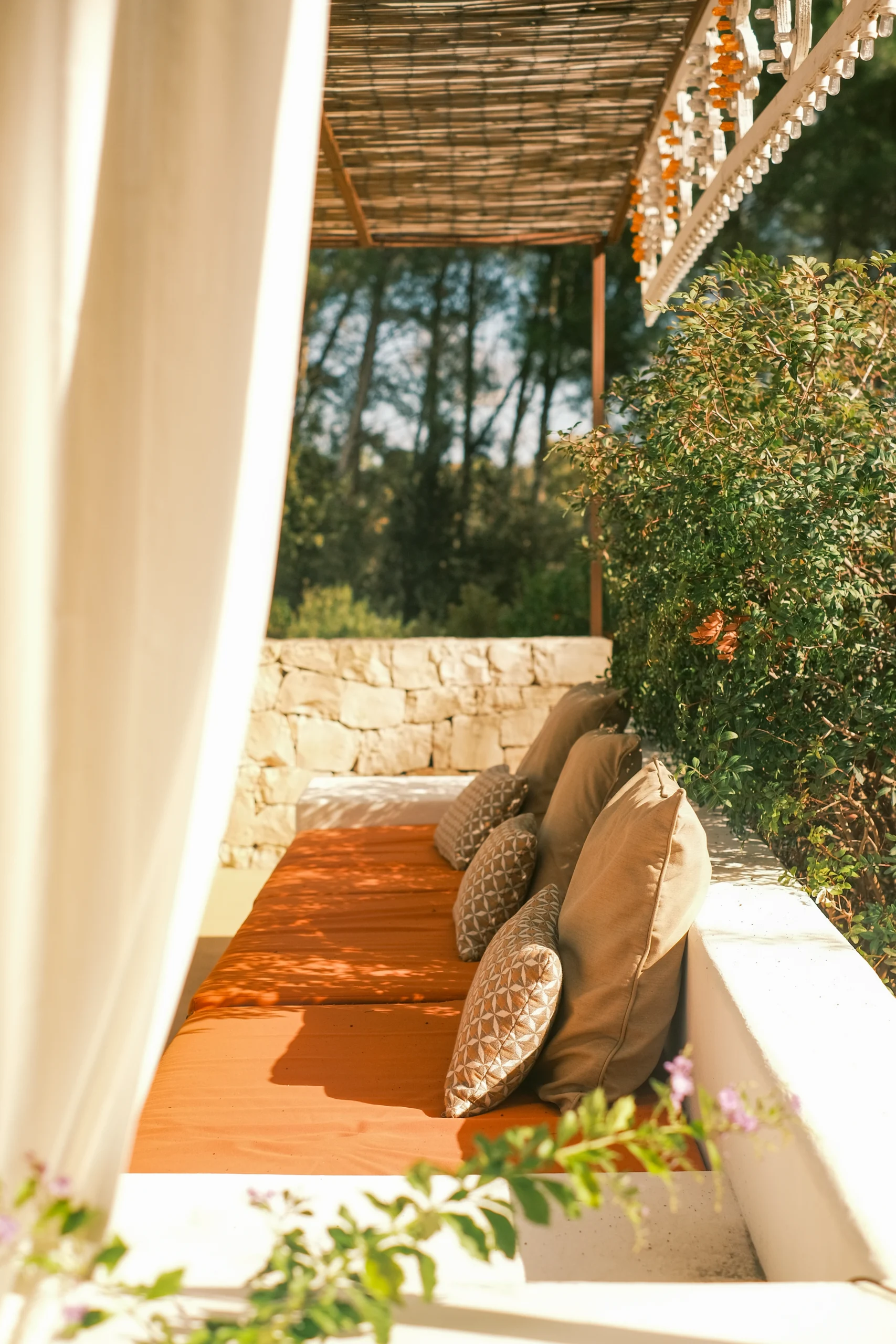 Outdoor lounge bed with terracotta cushions beneath a bamboo pergola, surrounded by Mediterranean vegetation