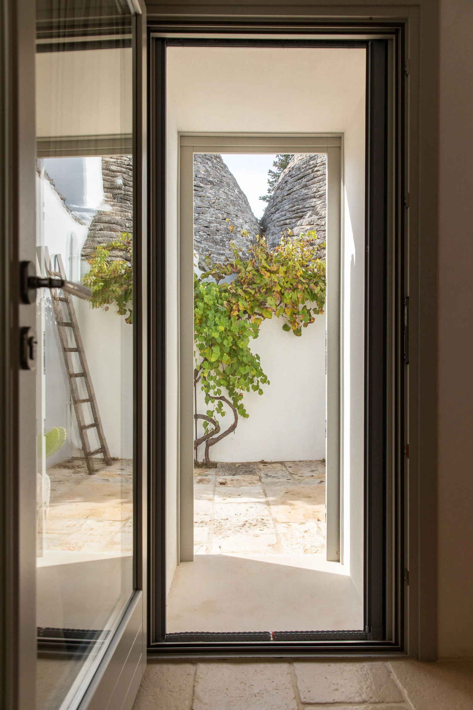 View from inside the villa through glass doors toward a trullo courtyard with stone walls and climbing vines