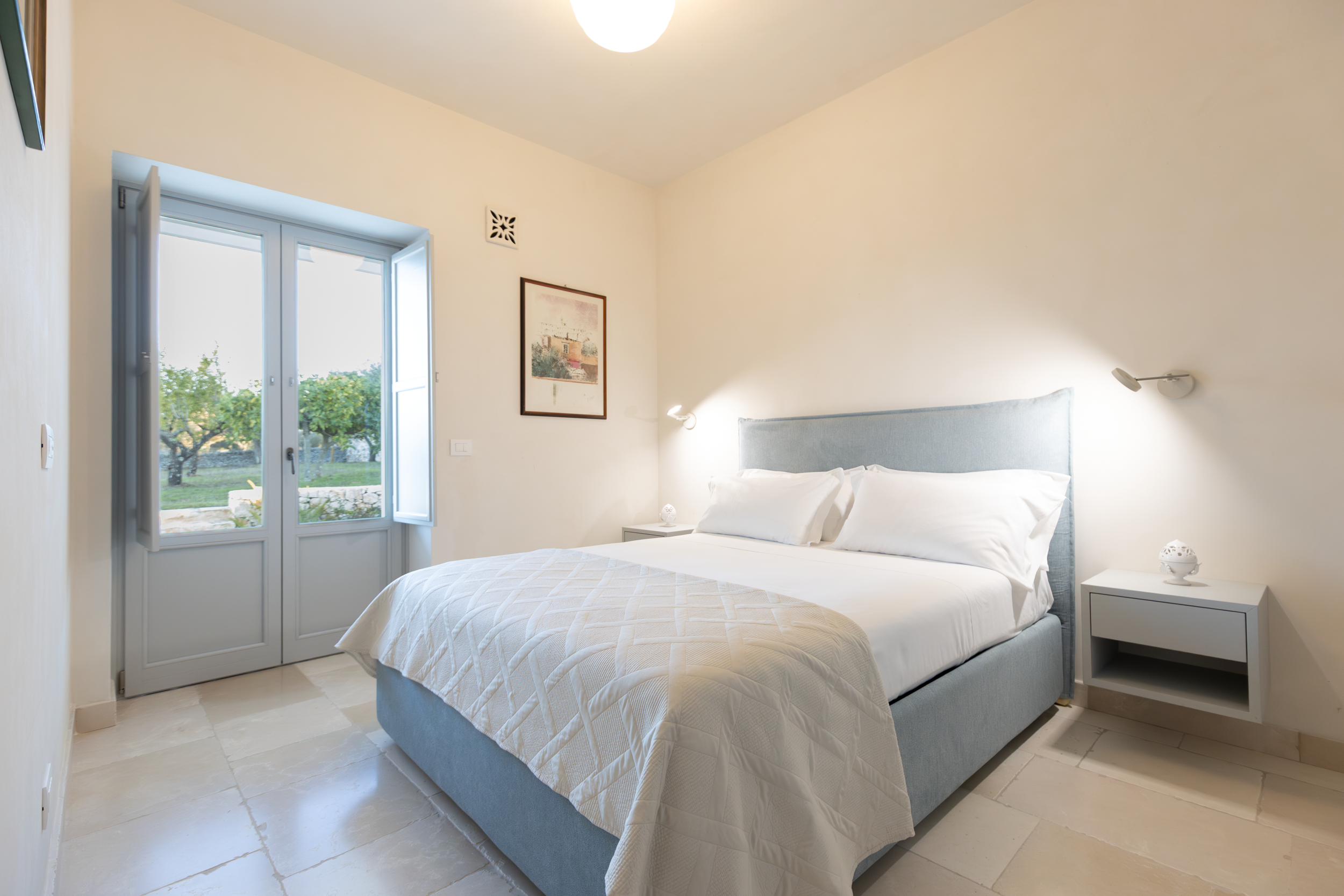 Bright master bedroom with blue upholstered bed, stone floor and garden view in a Puglia luxury villa