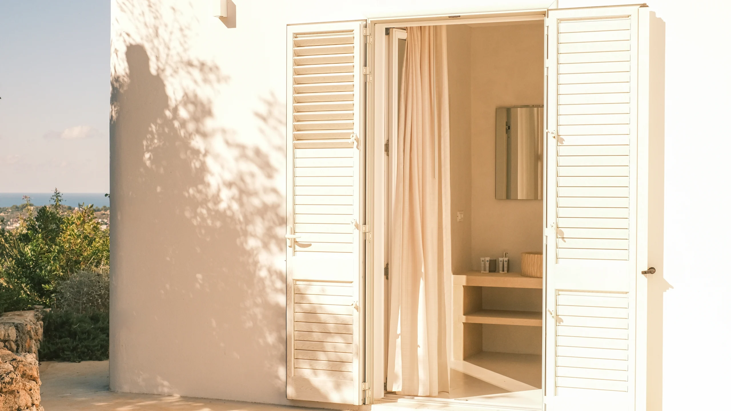 Villa Leucade outdoor bathroom entrance with white shutters, soft natural light and sea view atmosphere in Puglia.
