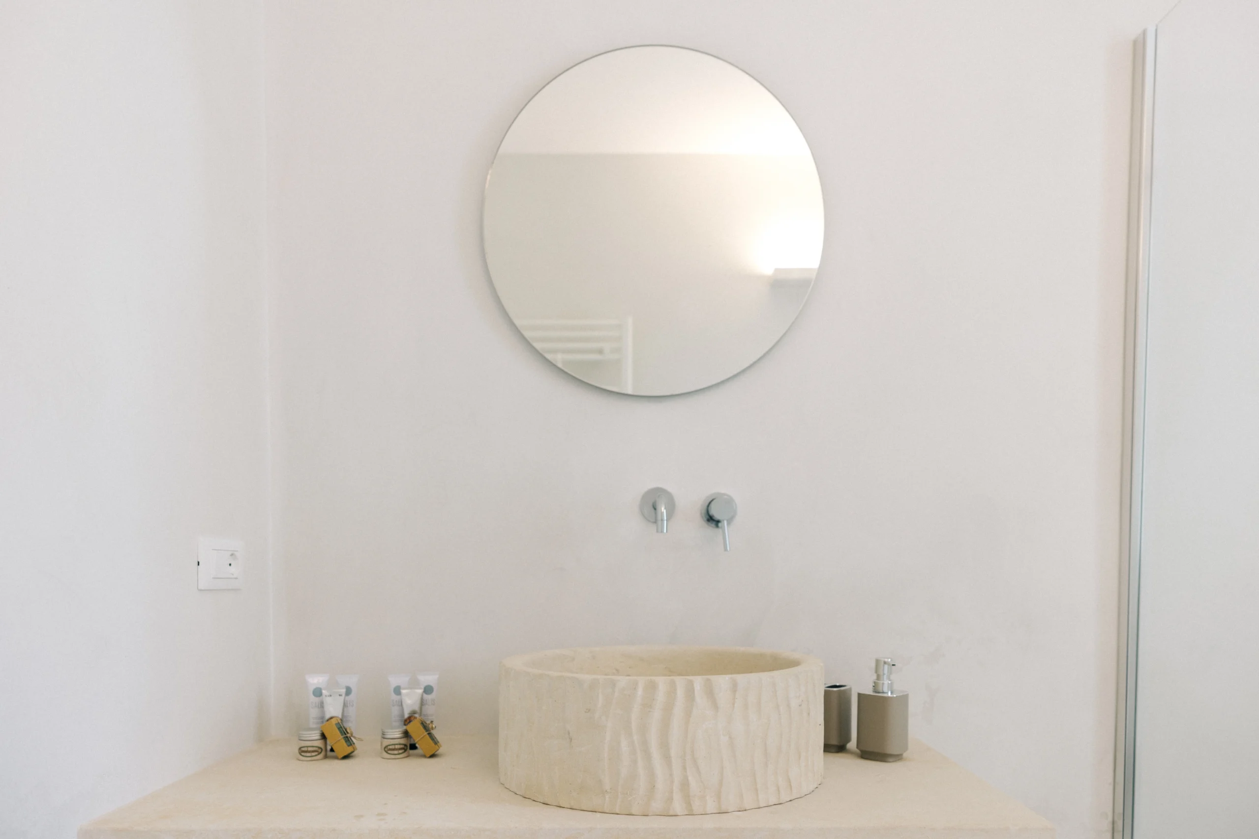 Villa Leucade round mirror above stone sink with elegant wall-mounted tap in luxury bathroom.