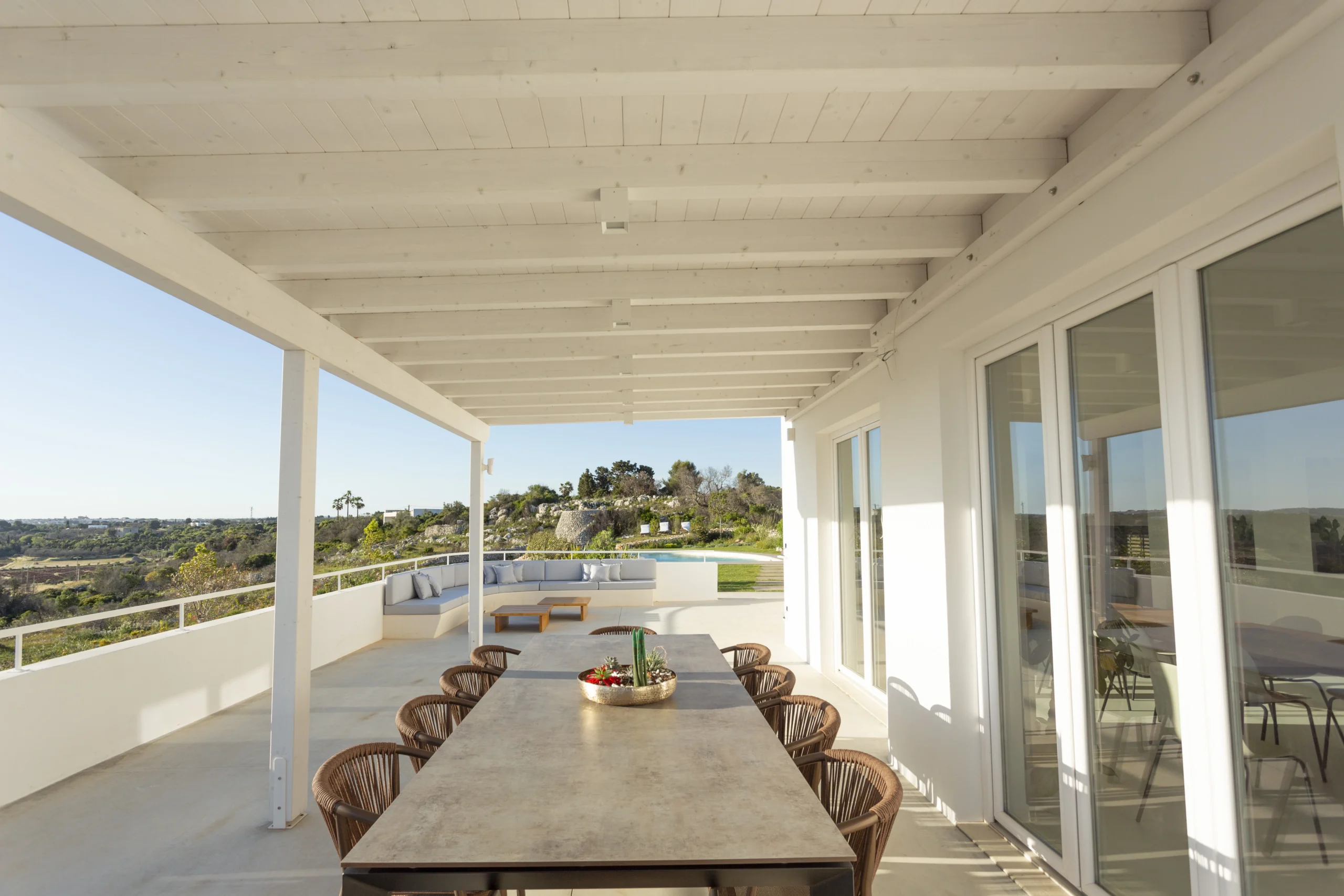 Covered terrace at Villa Leucade with long dining table, lounge seating and panoramic countryside views.