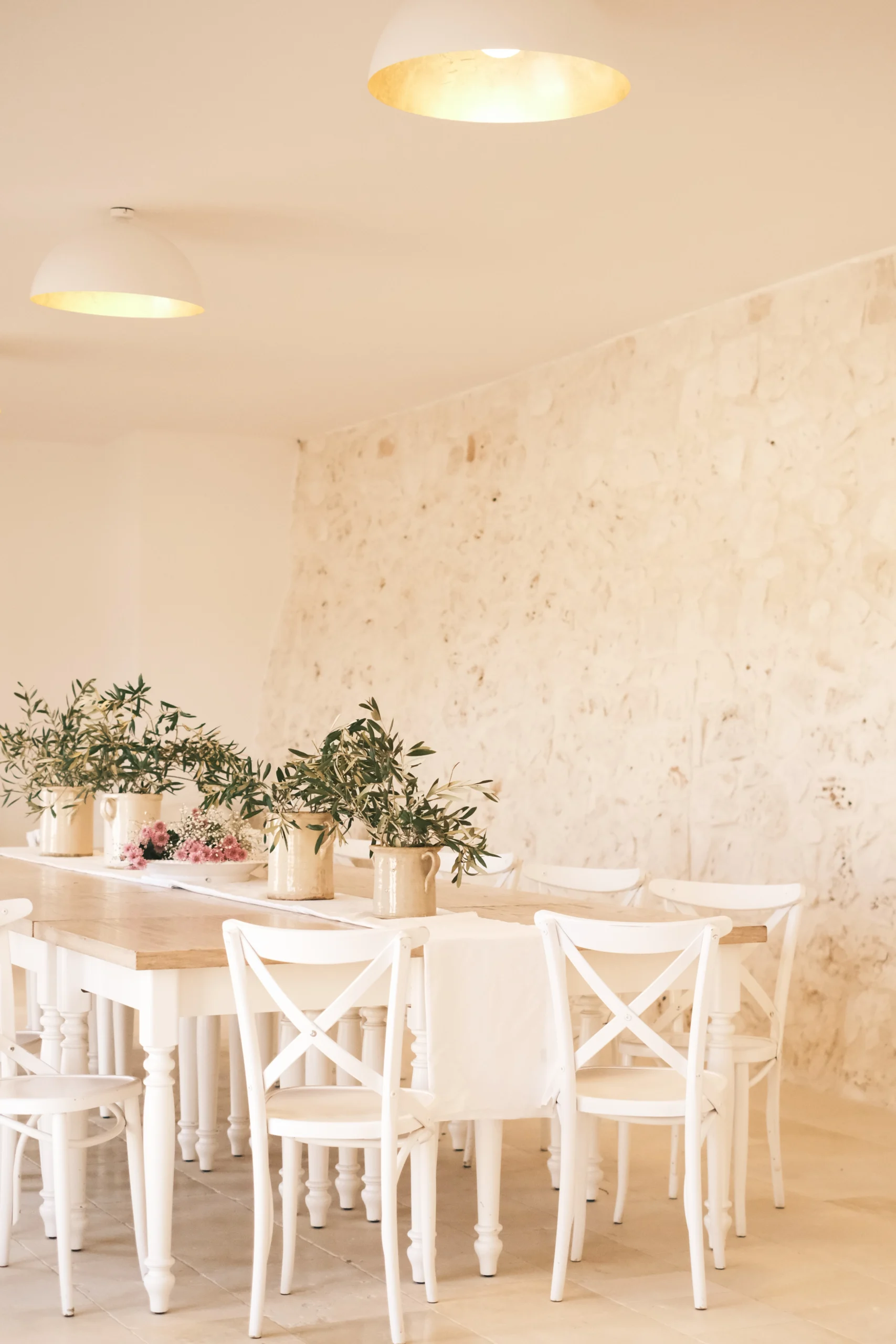 Elegant indoor dining area with long wooden table, white chairs, olive branch decor and natural stone walls in a luxury villa in Puglia