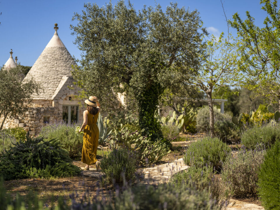Woman in yellow dress and straw hat walking along stone garden path lined with lavender, rosemary and cacti near a traditional trullo in Puglia