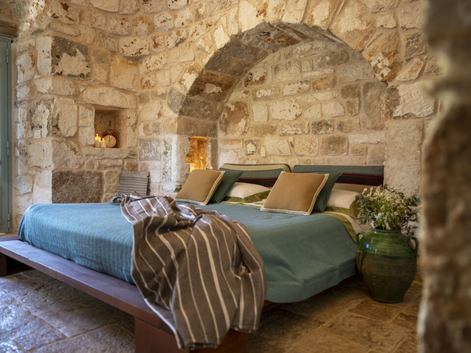 Rustic stone bedroom with vaulted arch, low wooden bed frame, sage green pillows, candles in wall niche and green ceramic vase inside a trullo in Puglia
