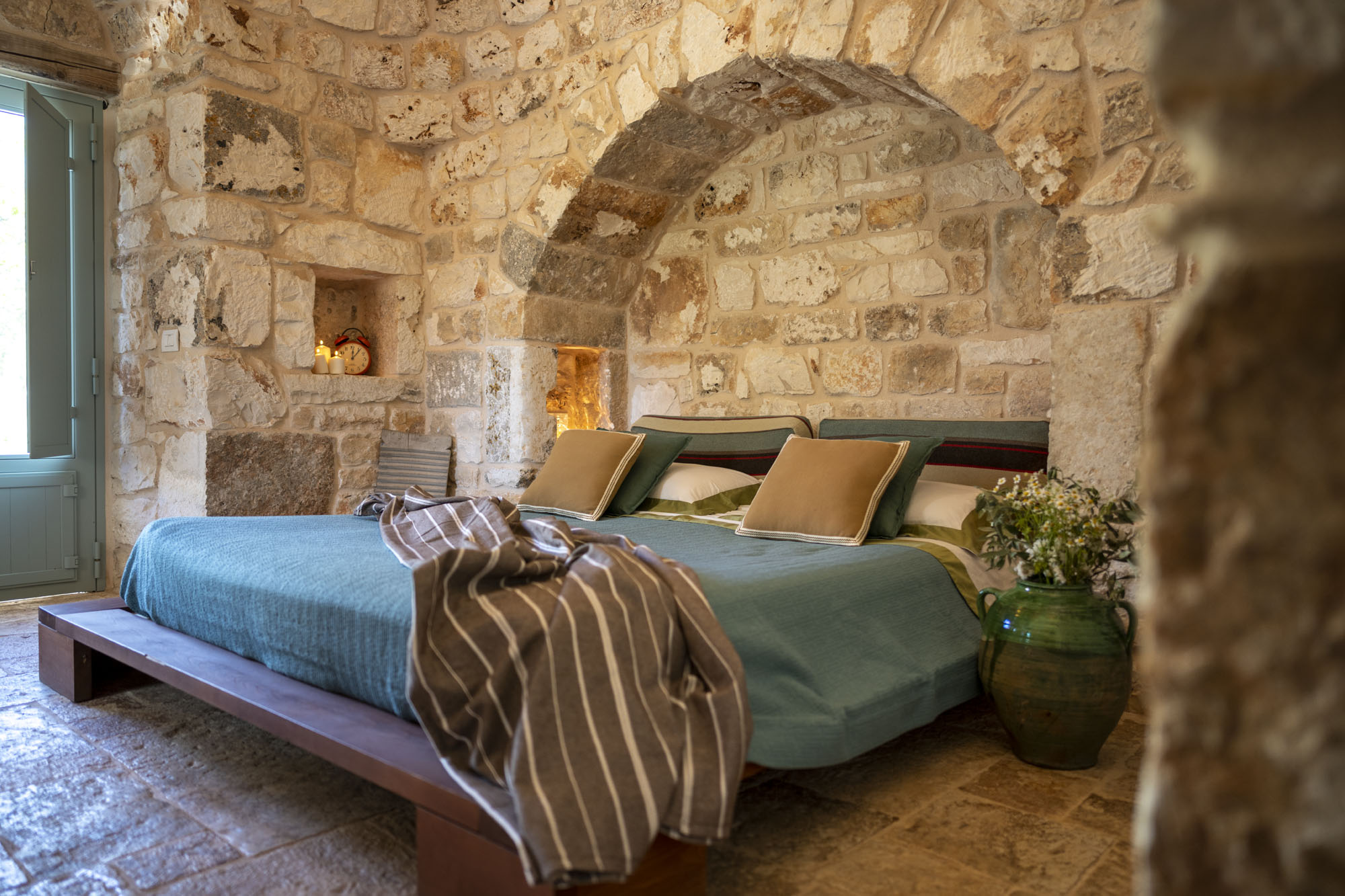 Rustic stone bedroom with vaulted arch, low wooden bed frame, sage green pillows, candles in wall niche and green ceramic vase inside a trullo in Puglia