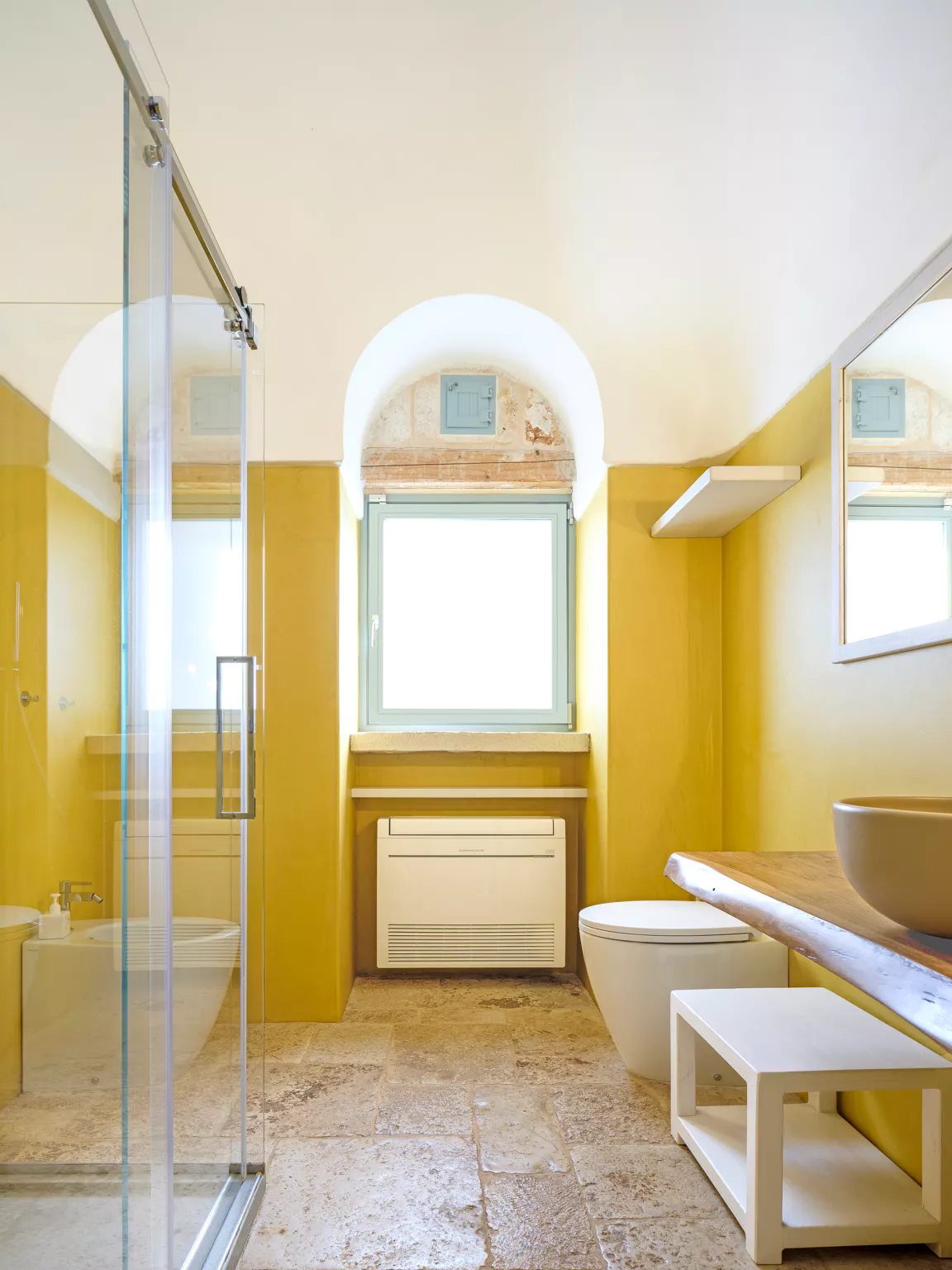 Full view of a yellow micro-cement en-suite bathroom with glass walk-in shower, stone vessel sink on live-edge wood shelf, arched plastered ceiling, sage blue shuttered window and travertine floor in a luxury Puglia villa