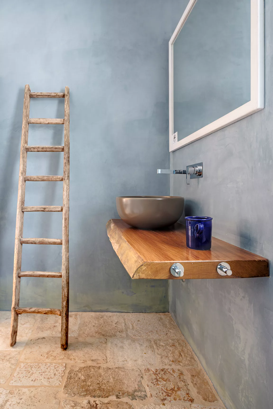 Minimalist bathroom with grey micro-cement walls, live-edge wood floating shelf, round stone vessel sink and vintage wooden ladder in a Puglia villa