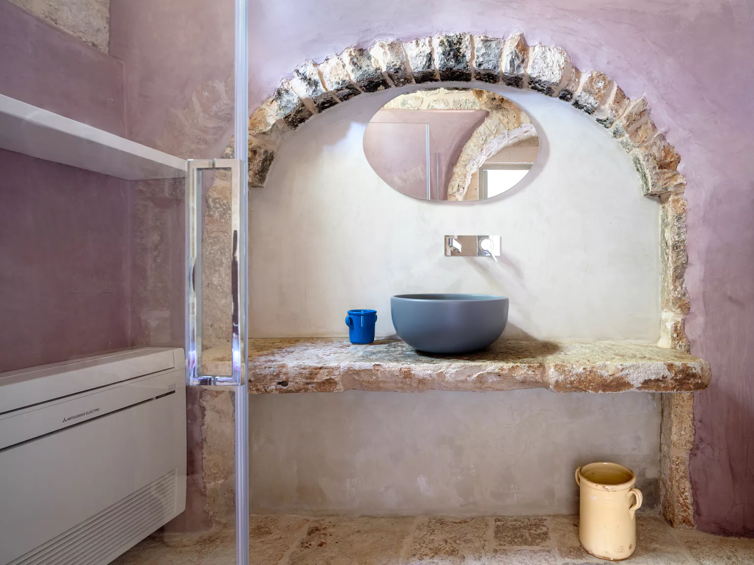 Puglia trullo bathroom with raw stone arch, blue ceramic vessel sink on ancient stone ledge, oval mirror and dusty rose micro-cement walls