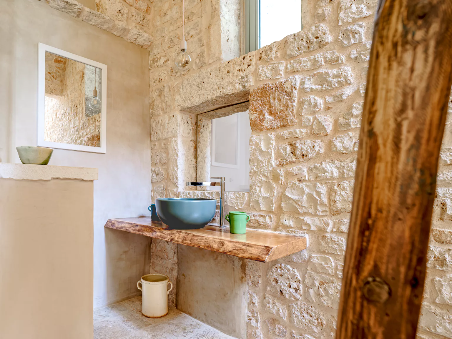 Bright bathroom in a Puglia stone villa with live-edge wood shelf, teal ceramic vessel sink, antique wooden door frame and exposed limestone walls