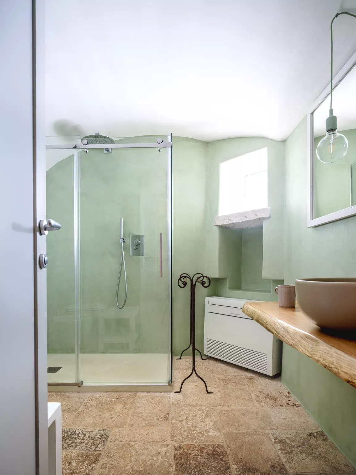 Soft sage green bathroom with vaulted plaster ceiling, glass-enclosed walk-in shower, live-edge wood vanity shelf, globe pendant light and travertine floor in a Puglia villa