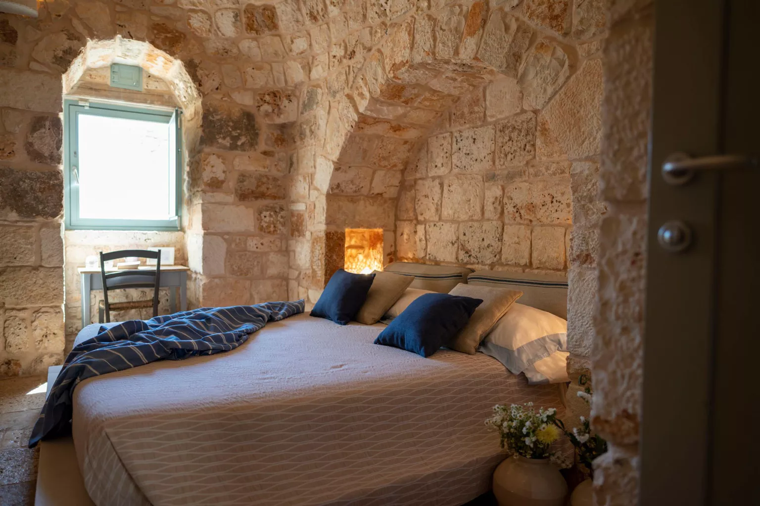 Intimate trullo bedroom with rough limestone walls, stone arched window, Himalayan salt lamp headboard niche, navy blue pillows and wildflower bouquet in Puglia