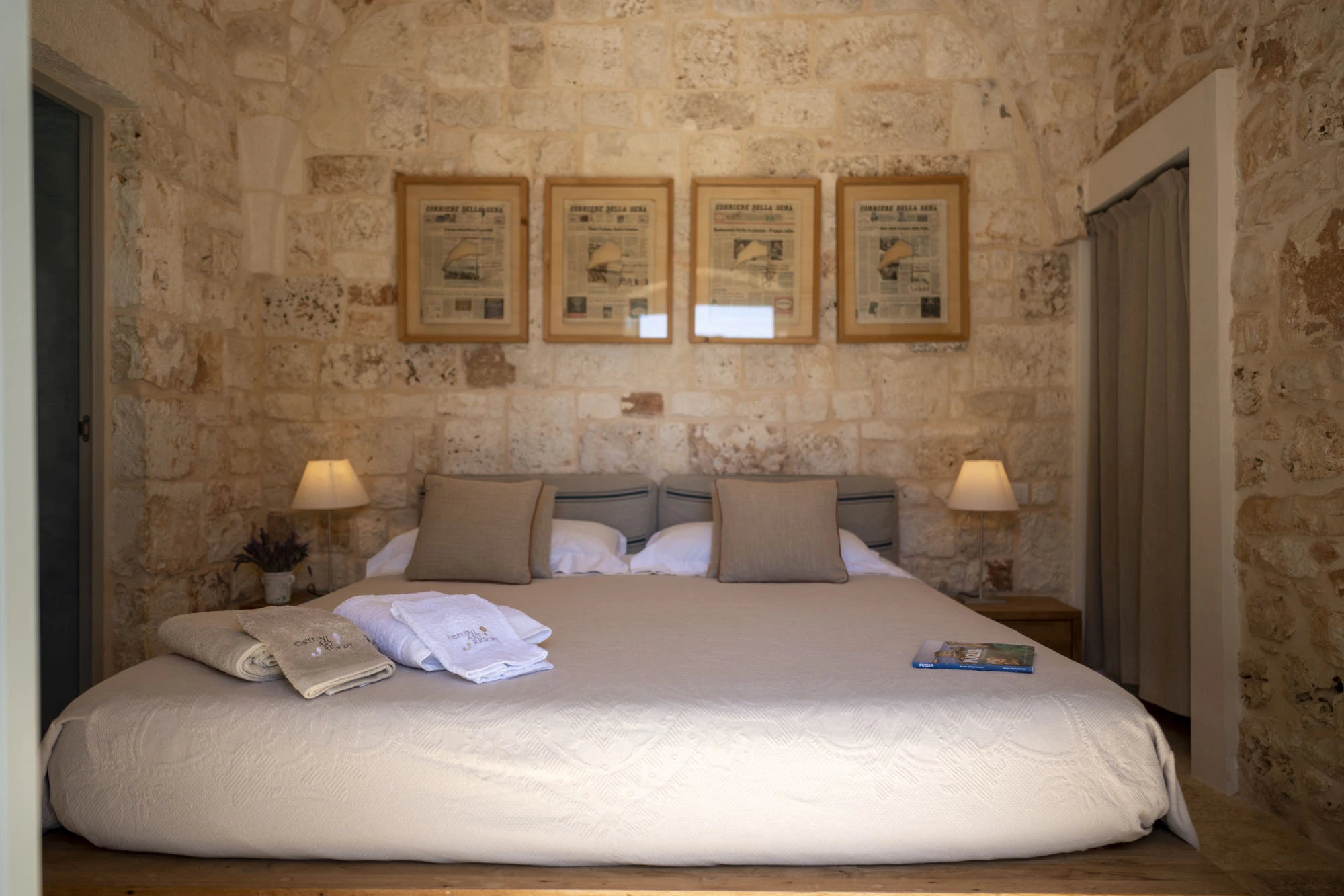 Elegant stone bedroom with four framed Corriere della Sera newspaper front pages above a double bed with taupe cushions and bedside lamps in a Puglia villa