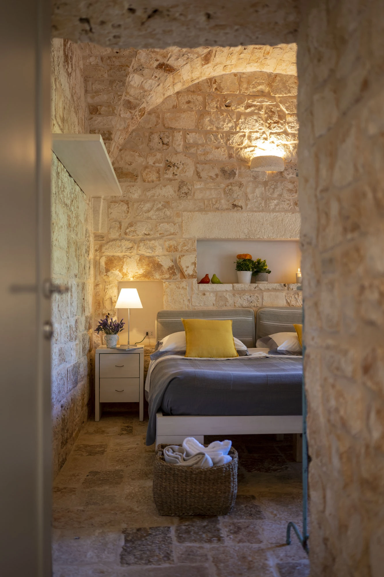 Cosy double bedroom viewed through a stone arched doorway at dusk, with yellow cushion, bedside lamp, lavender bouquet and wicker towel basket in a Puglia villa