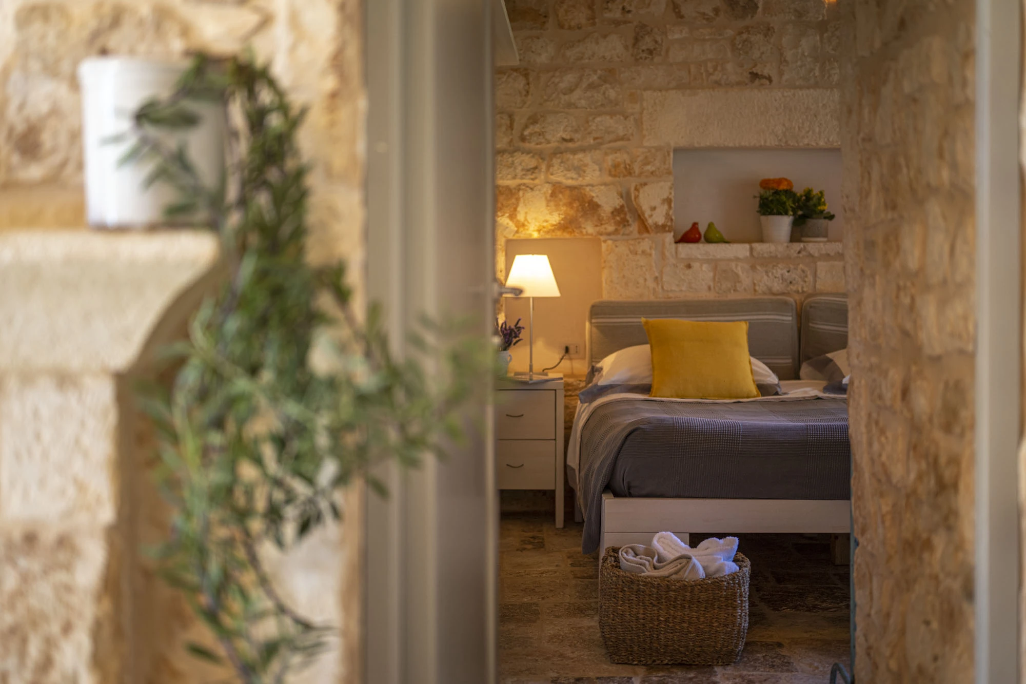 Double bedroom in a Puglia stone villa seen through a stone arch with a cascading olive branch in foreground, warm lamp light, yellow cushion and wicker basket