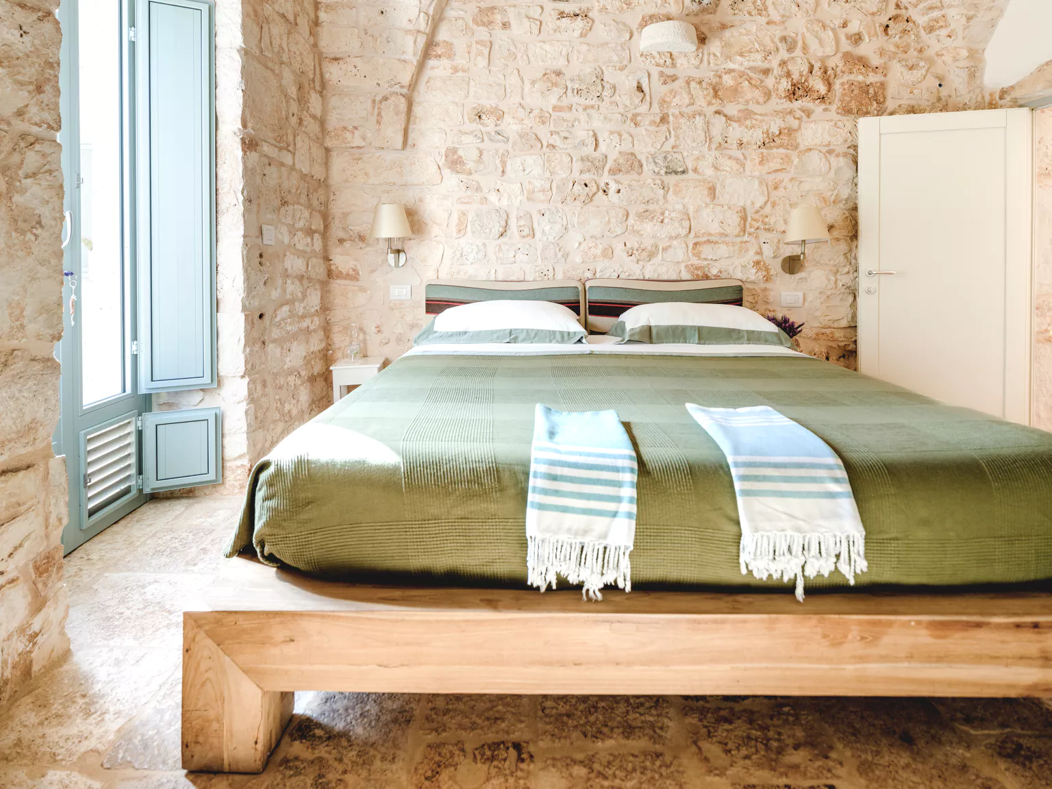 Bright double bedroom with exposed limestone walls, olive green bedspread, solid wood bed frame, striped fouta towels and sage blue shuttered door in a Puglia villa
