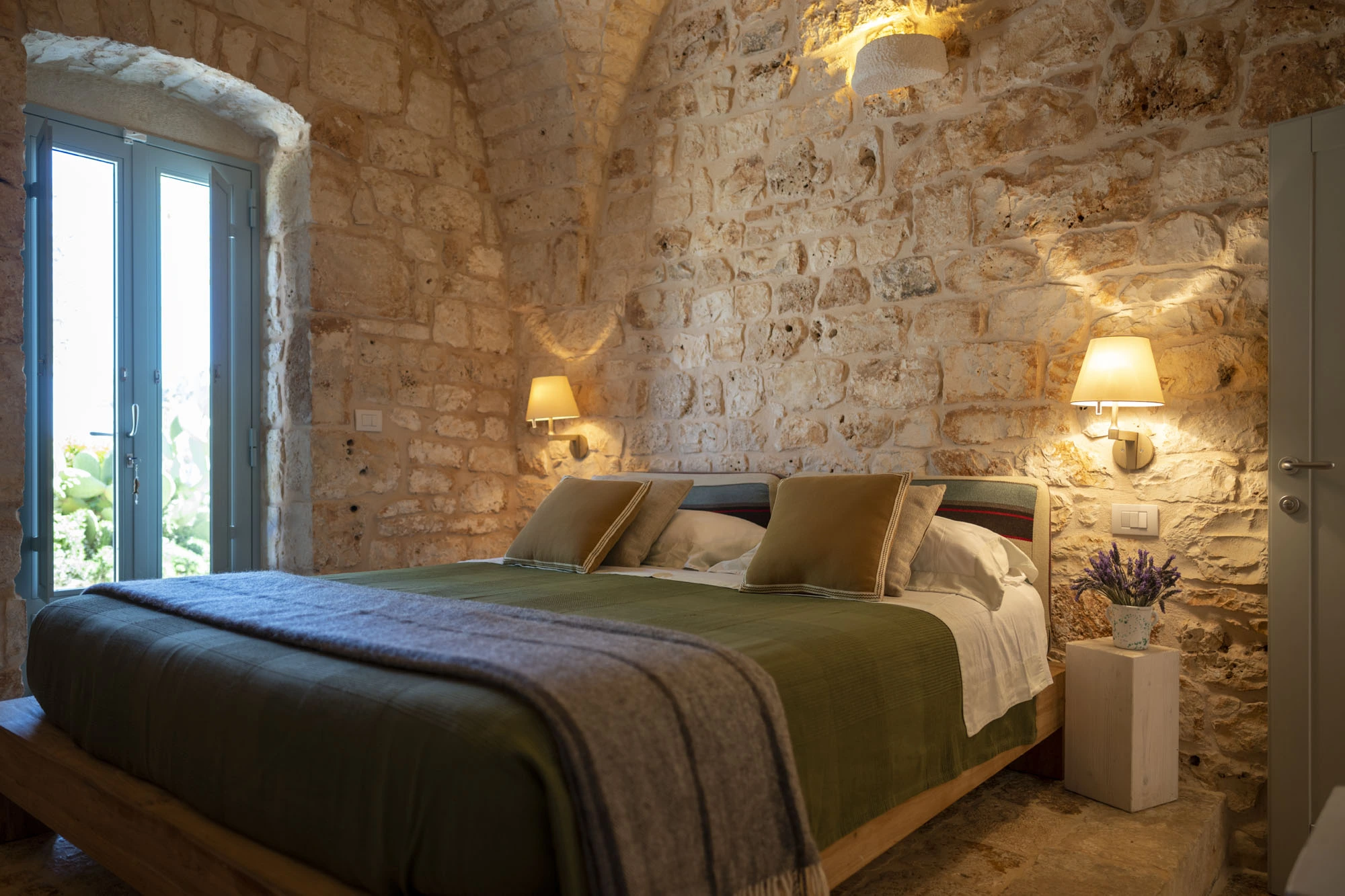 Romantic double bedroom with vaulted limestone ceiling, olive green bedding, earth-toned cushions, wall-mounted bedside lamps and lavender bouquet in a Puglia countryside villa