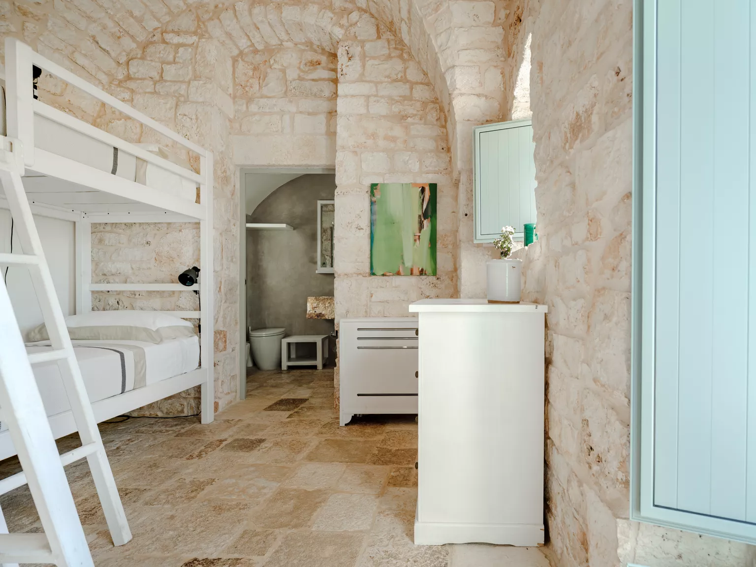 Family bedroom with white bunk beds, exposed limestone stone walls, arched doorway opening to en-suite bathroom with raw stone sink in a Puglia countryside villa