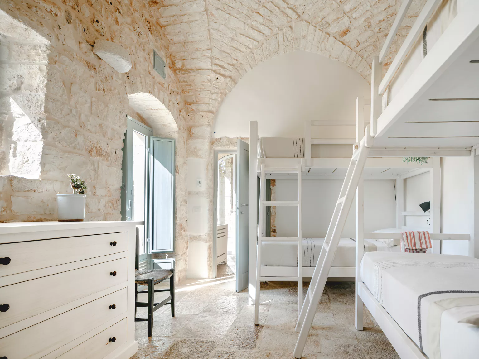 Bright vaulted bunk bedroom with white bunk beds, white dresser, ancient stone ceiling arches, sage blue shutters and travertine floor in a luxury Puglia villa