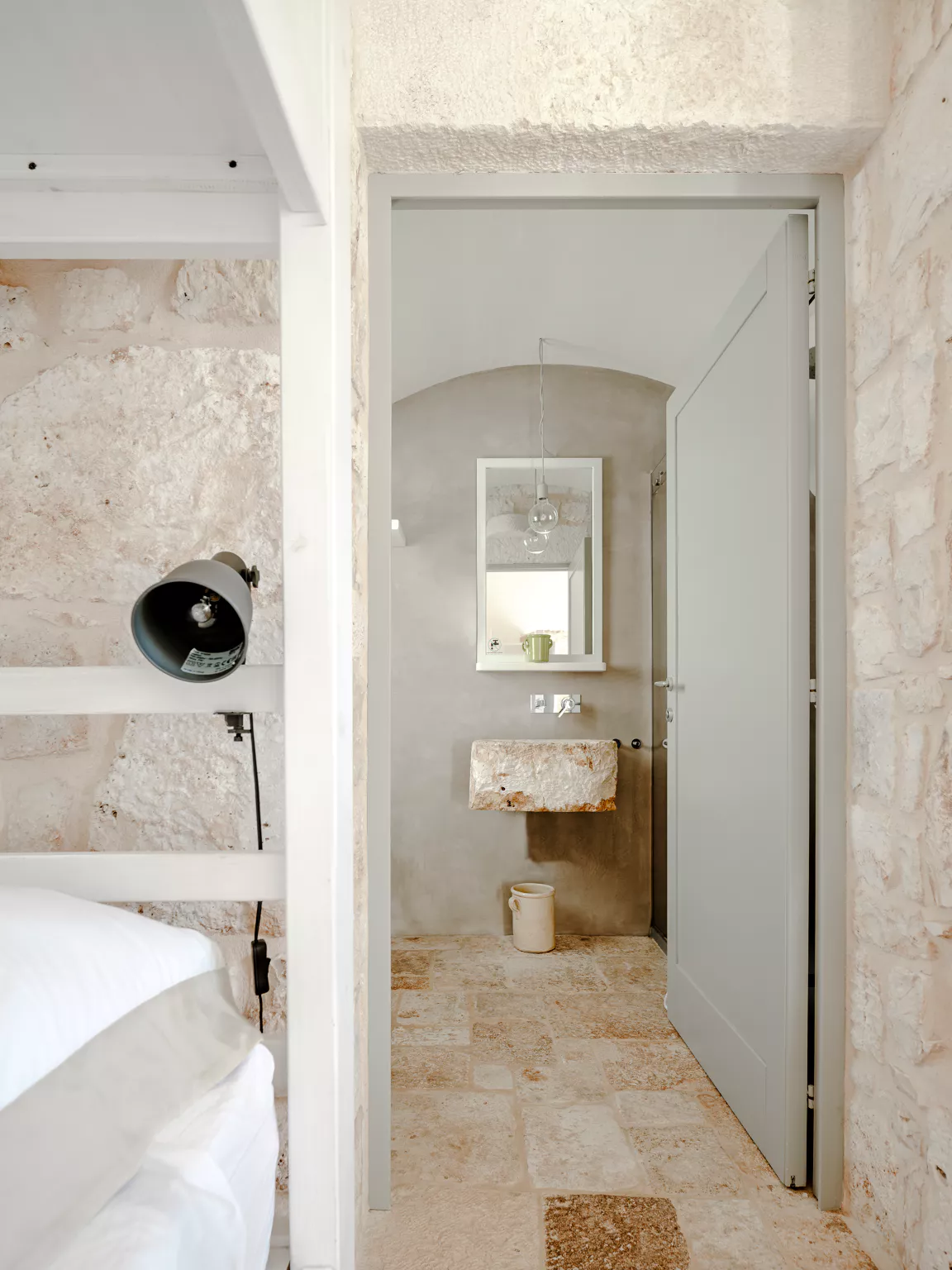 Minimalist en-suite bathroom adjacent to bunk bedroom, featuring a carved stone sink basin, grey micro-cement walls, globe pendant light and arched doorway in a Puglia villa