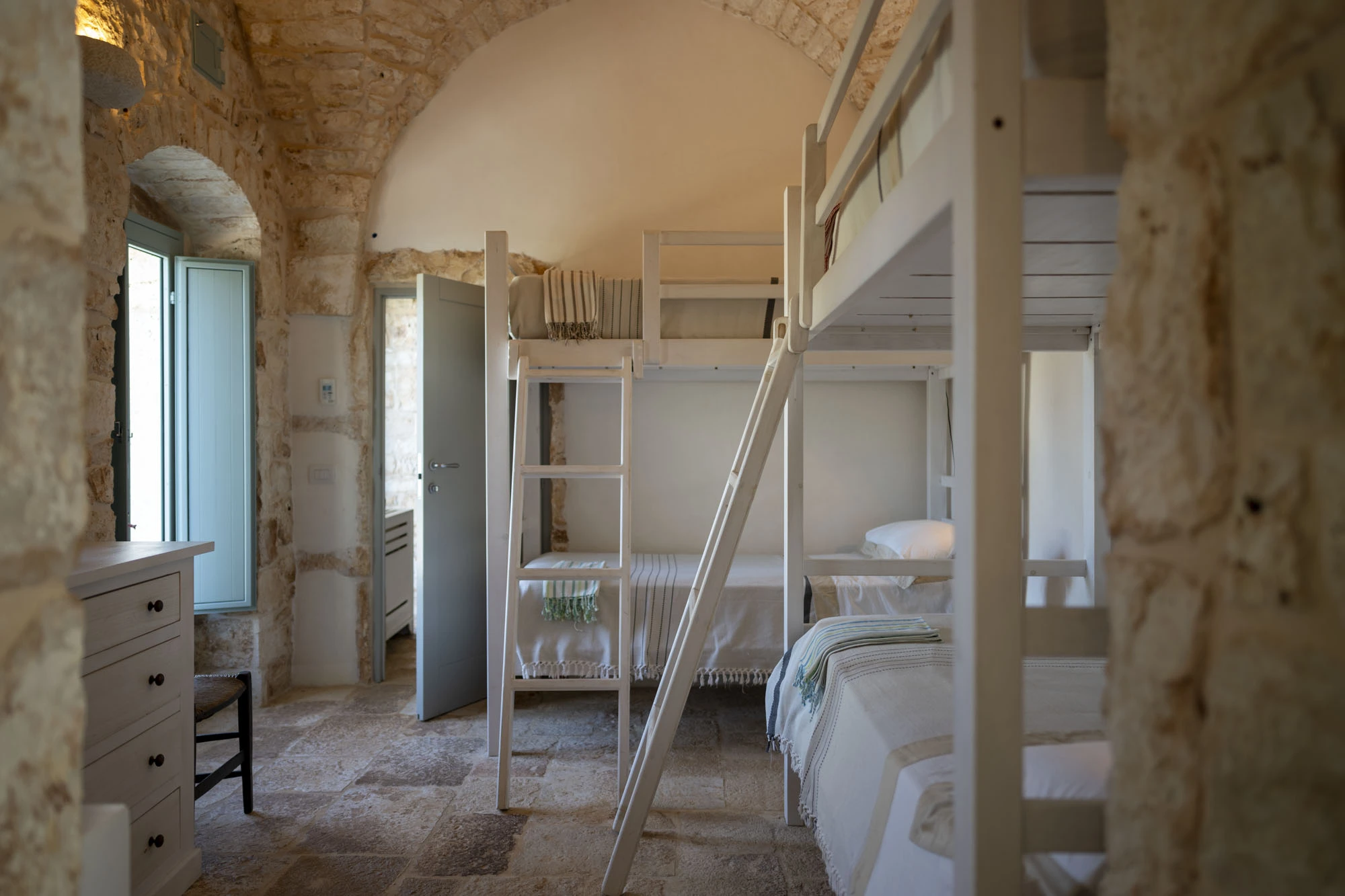 Airy family bunk bedroom with vaulted stone arch, two white bunk beds with striped textile throws, white dresser and sage shutters in a luxury Puglia villa