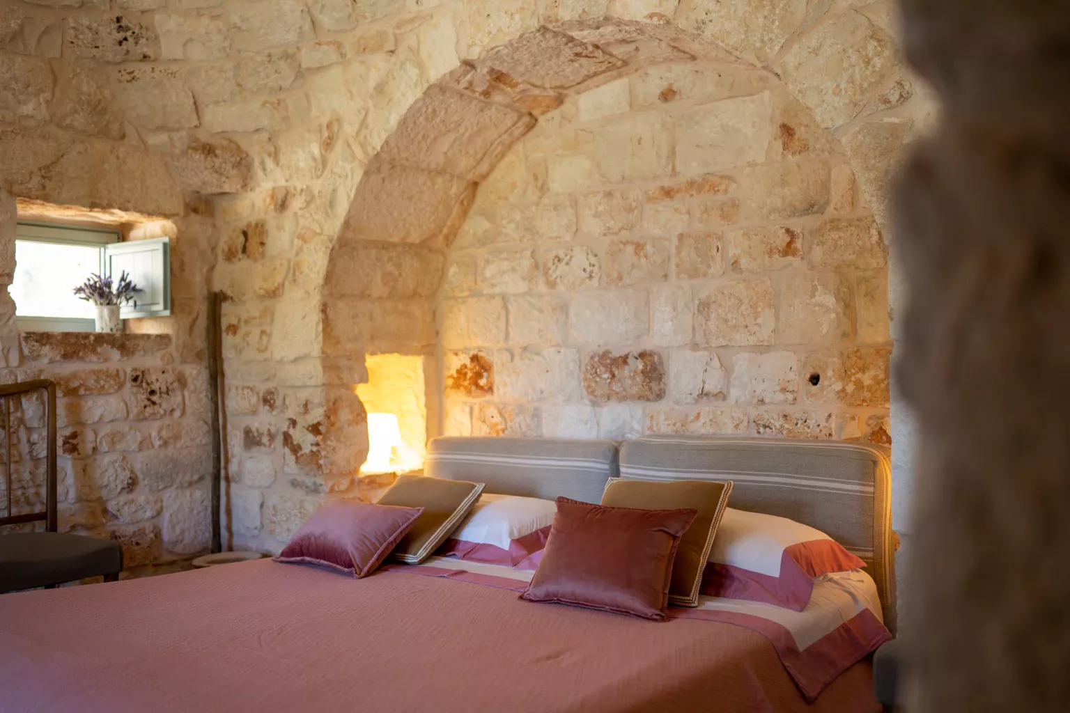 Romantic double bedroom inside a trullo with stone arch headboard niche, rose pink bedspread, dusty pink velvet cushions, backlit stone wall and lavender on the windowsill in Puglia