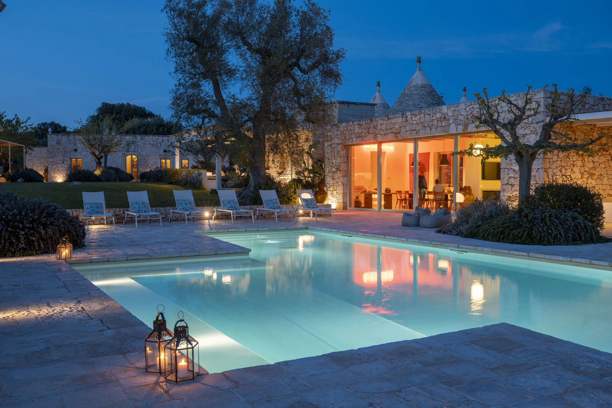 Luxury Puglia trulli villa at night with glowing swimming pool, Moroccan lanterns on terrace, lit interior through sliding glass doors, ancient olive trees and stone walls under a blue dusk sky