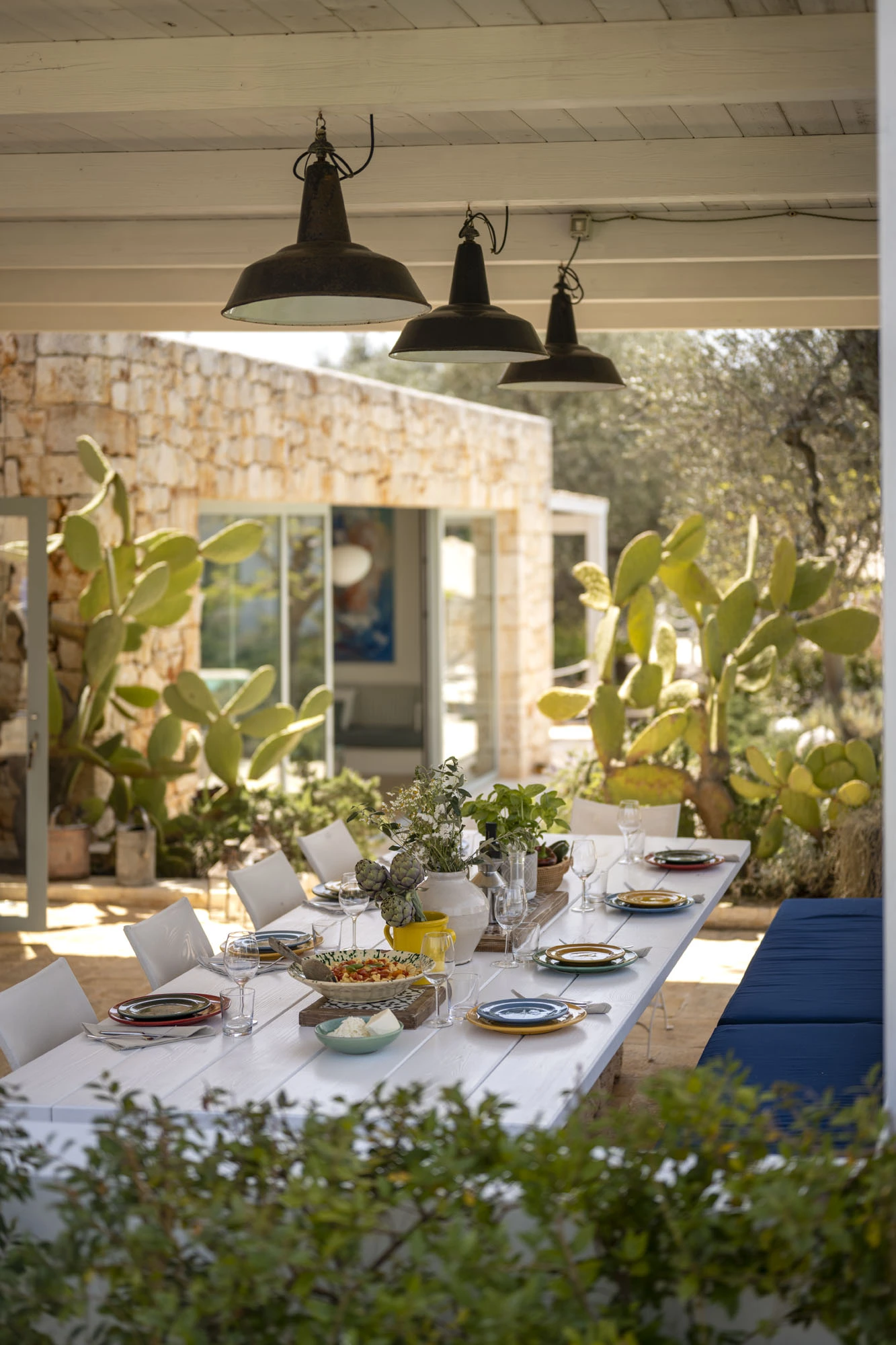 Long white outdoor dining table set for lunch under a pergola with industrial pendant lamps, colorful ceramic plates, fresh artichokes and herbs, surrounded by prickly pear cacti and olive trees at a Puglia villa