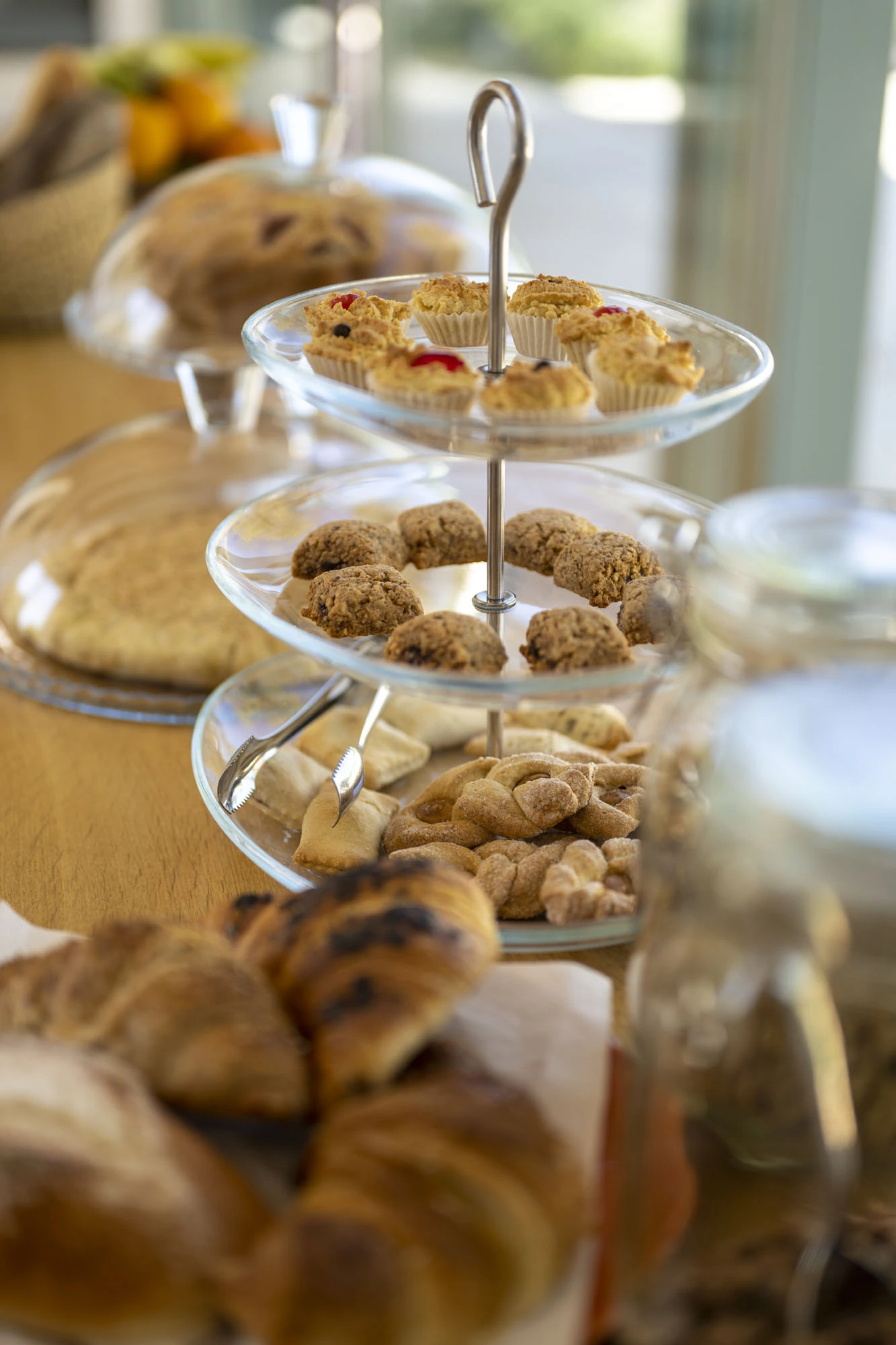 Three-tier glass cake stand with homemade Apulian cookies, taralli, fruit tarts and mini muffins alongside chocolate croissants and artisanal breads on a villa breakfast table in Puglia