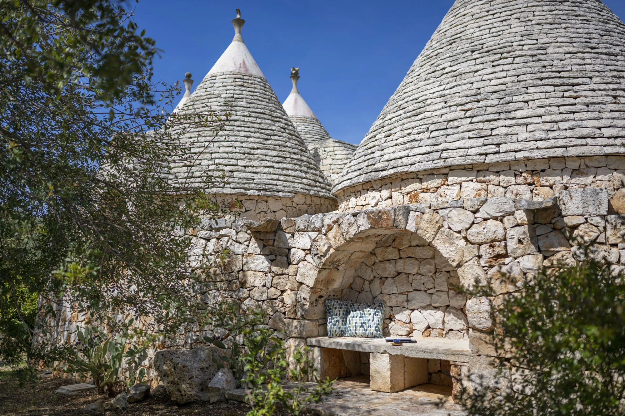 Carved stone bench set into a dry-stone arched wall niche with printed cushion, surrounded by trulli cones with traditional grey stone roofs and olive branches in a Puglia villa garden