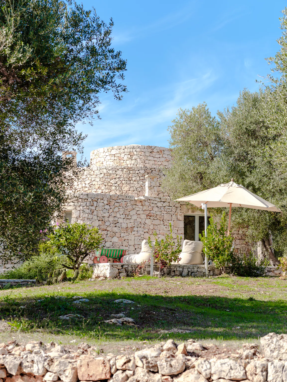Restored circular stone tower villa with cream parasol, outdoor bean bag chairs and garden bench amid olive trees and flowering shrubs in the Puglia countryside
