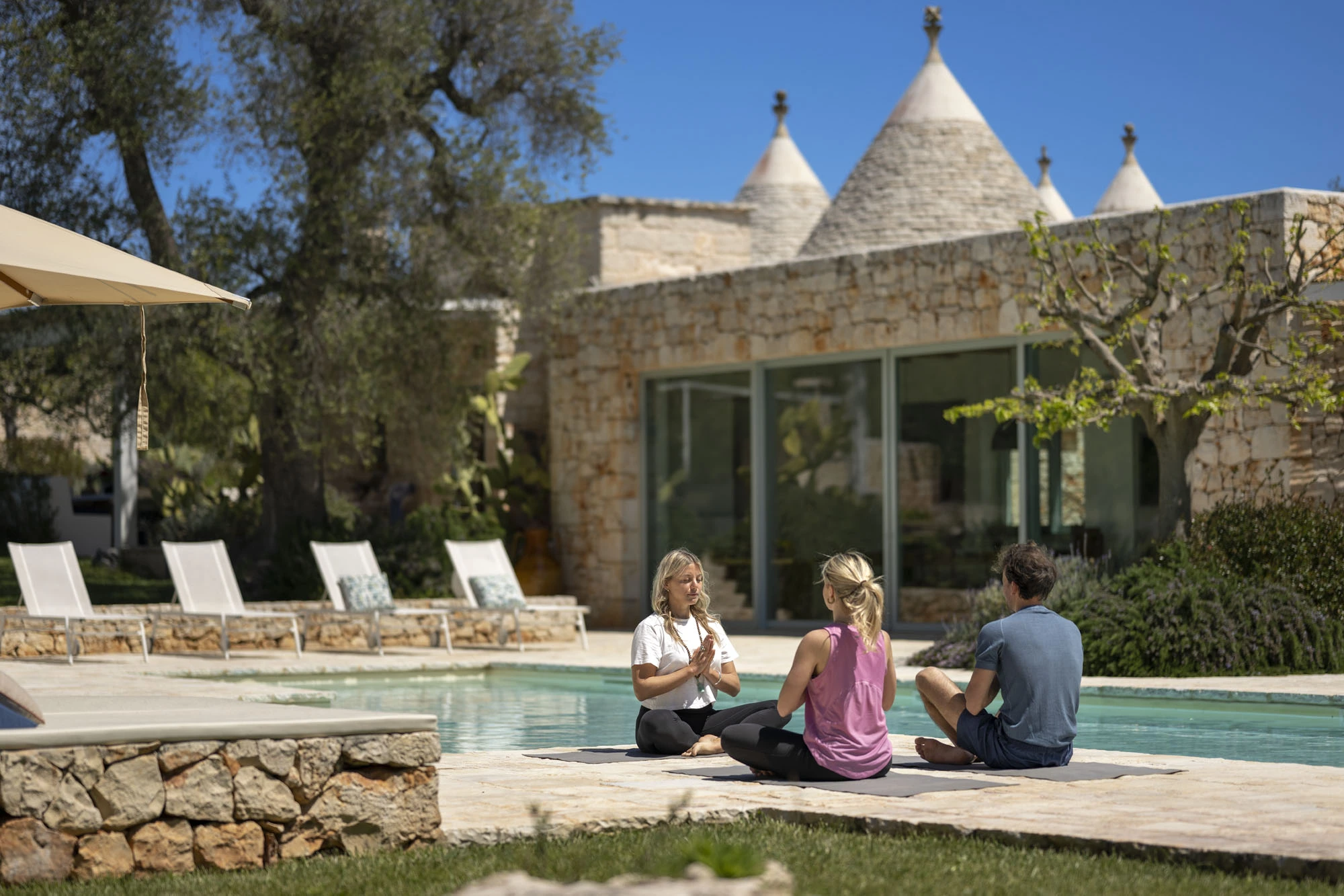 Three people in a yoga session beside a swimming pool at a luxury trulli villa in Puglia, with stone architecture and olive trees in the background under a blue sky