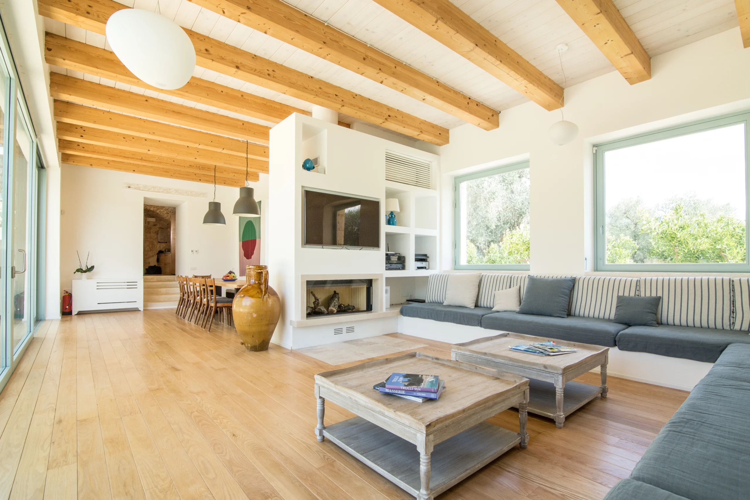Spacious open-plan living room with exposed wood beam ceiling, large corner grey sofa, double wooden coffee tables, fireplace, TV wall unit and dining area with olive tree views at a luxury Puglia villa