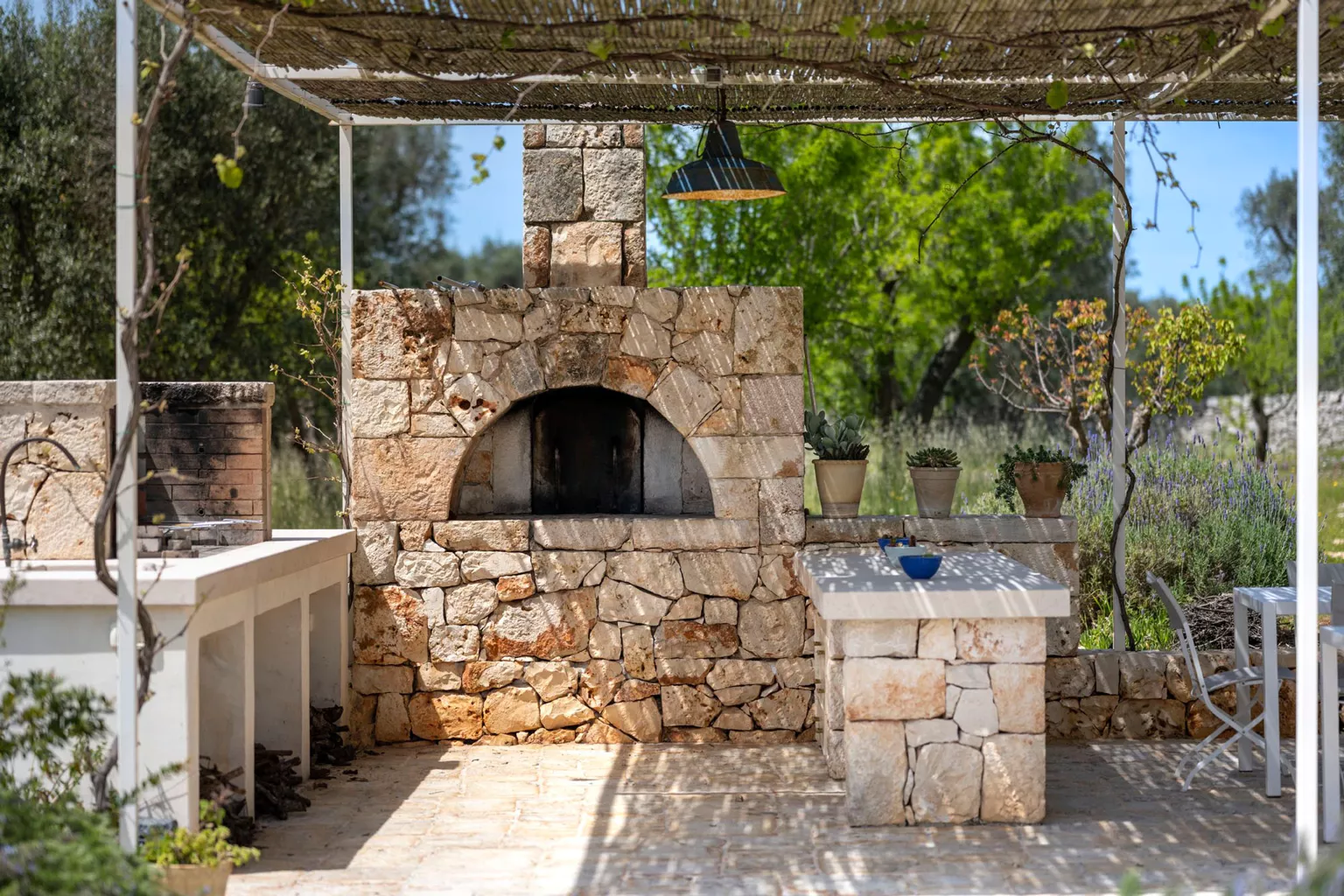 Hand-built stone wood-fired pizza oven with arched opening under a bamboo pergola with pendant lamp, stone prep counters and potted succulents at a luxury Puglia villa