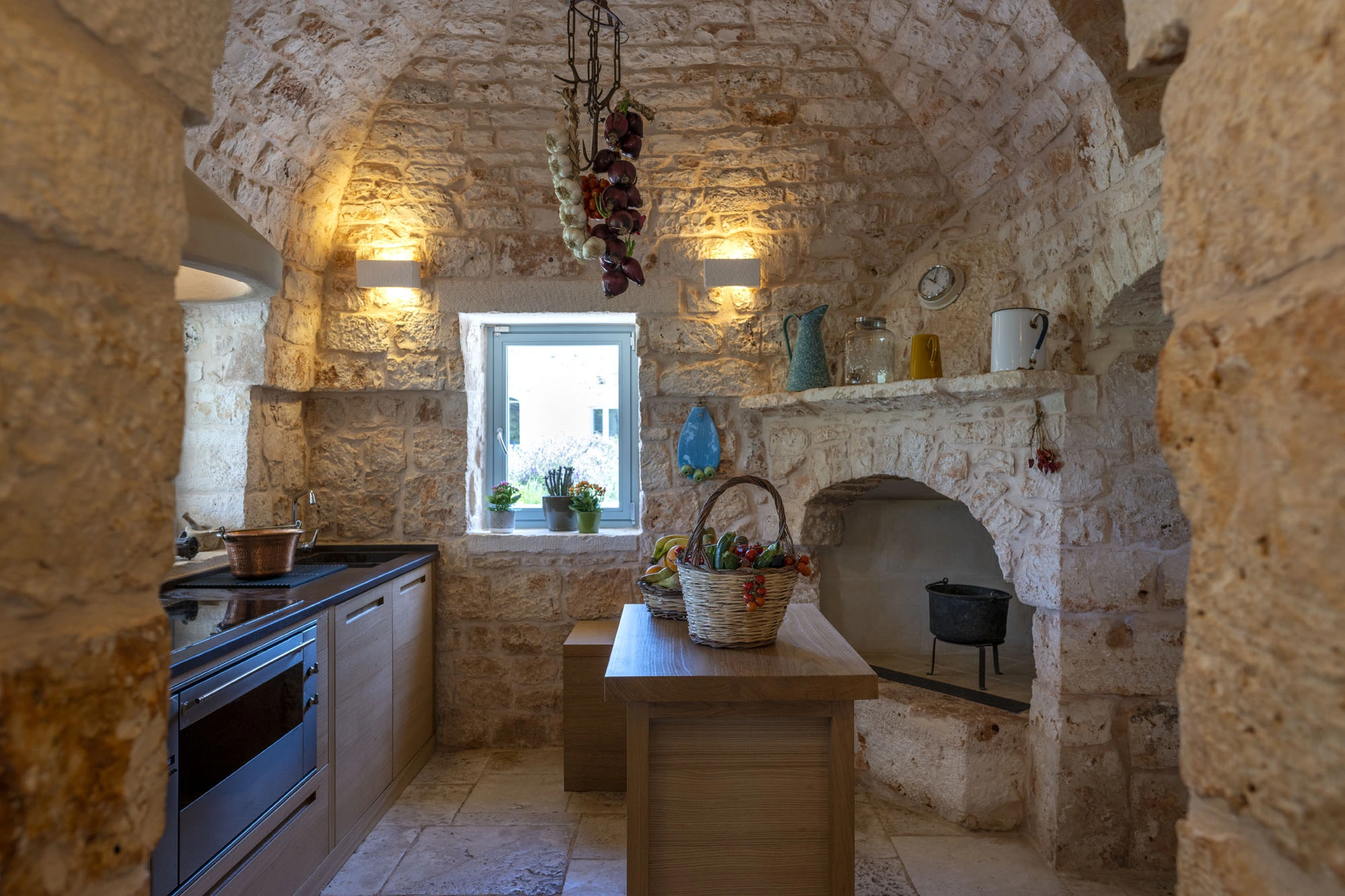 Atmospheric trullo kitchen with vaulted limestone ceiling, modern range cooker, wood island with fresh fruit basket, ancient fireplace niche and hanging onions and garlic in Puglia