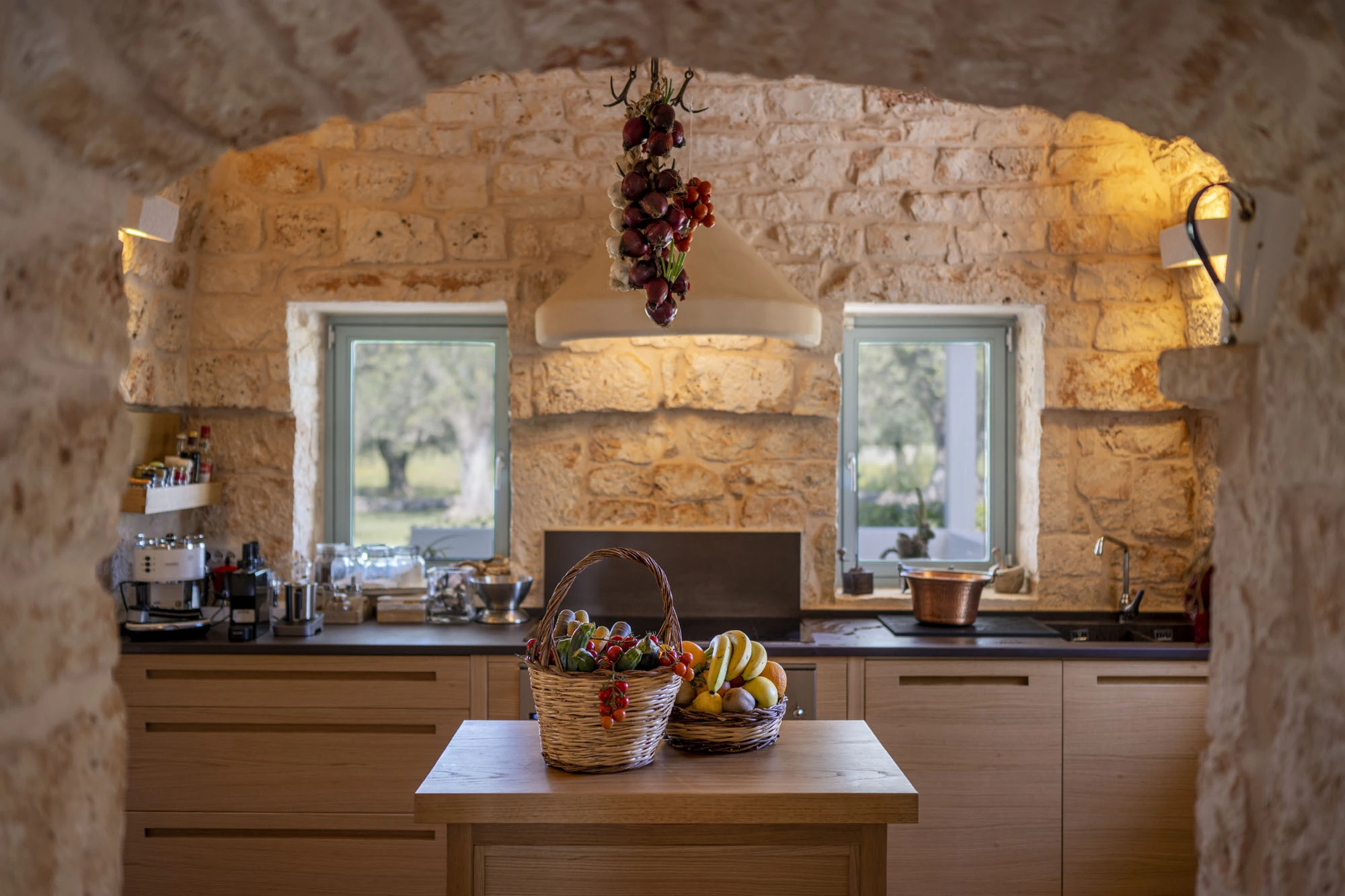 Stone arch framed kitchen interior with wood island unit, two wicker fruit baskets, dark stone countertops, oak cabinets and hanging red onions at a luxury Puglia trulli villa