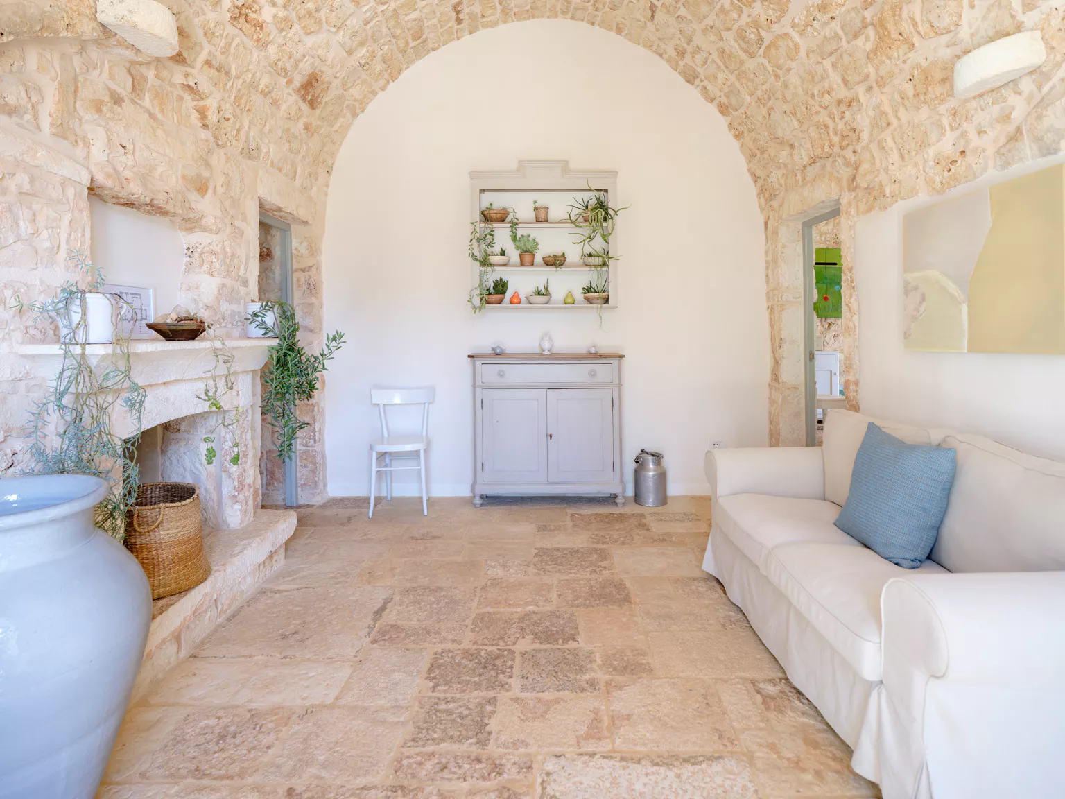 Bright stone vaulted living room inside a restored Puglian lamia with arched alcove, succulent plant shelf, grey dresser, white sofa with blue cushion and ancient limestone floor