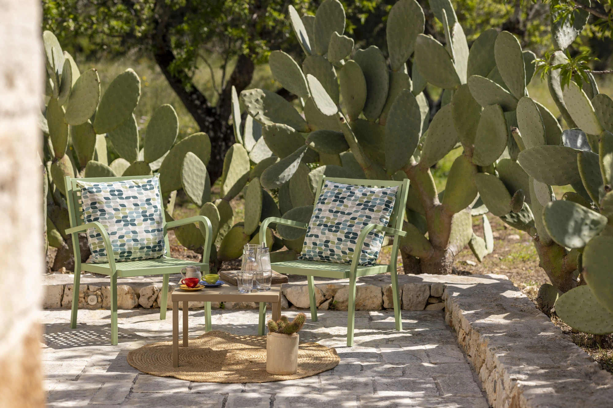 Two sage green outdoor armchairs with patterned cushions on a limestone terrace surrounded by large prickly pear cacti, with colourful ceramic cups and water glasses on a small wooden table at a Puglia villa