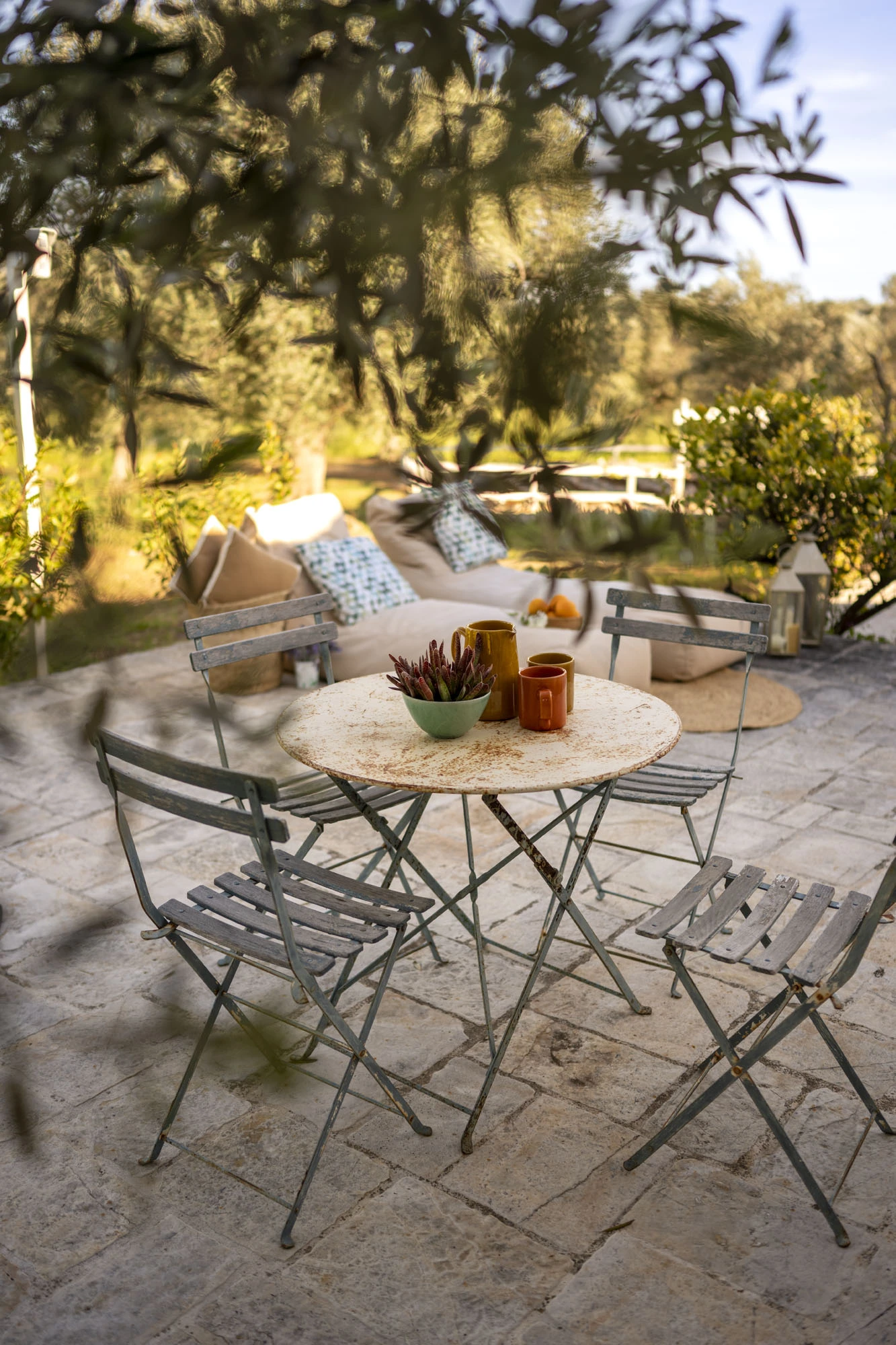 Weathered sage green bistro table and folding chairs with ceramic mugs on a limestone terrace at golden hour, with outdoor sofa, garden lantern and olive trees at a Puglia villa