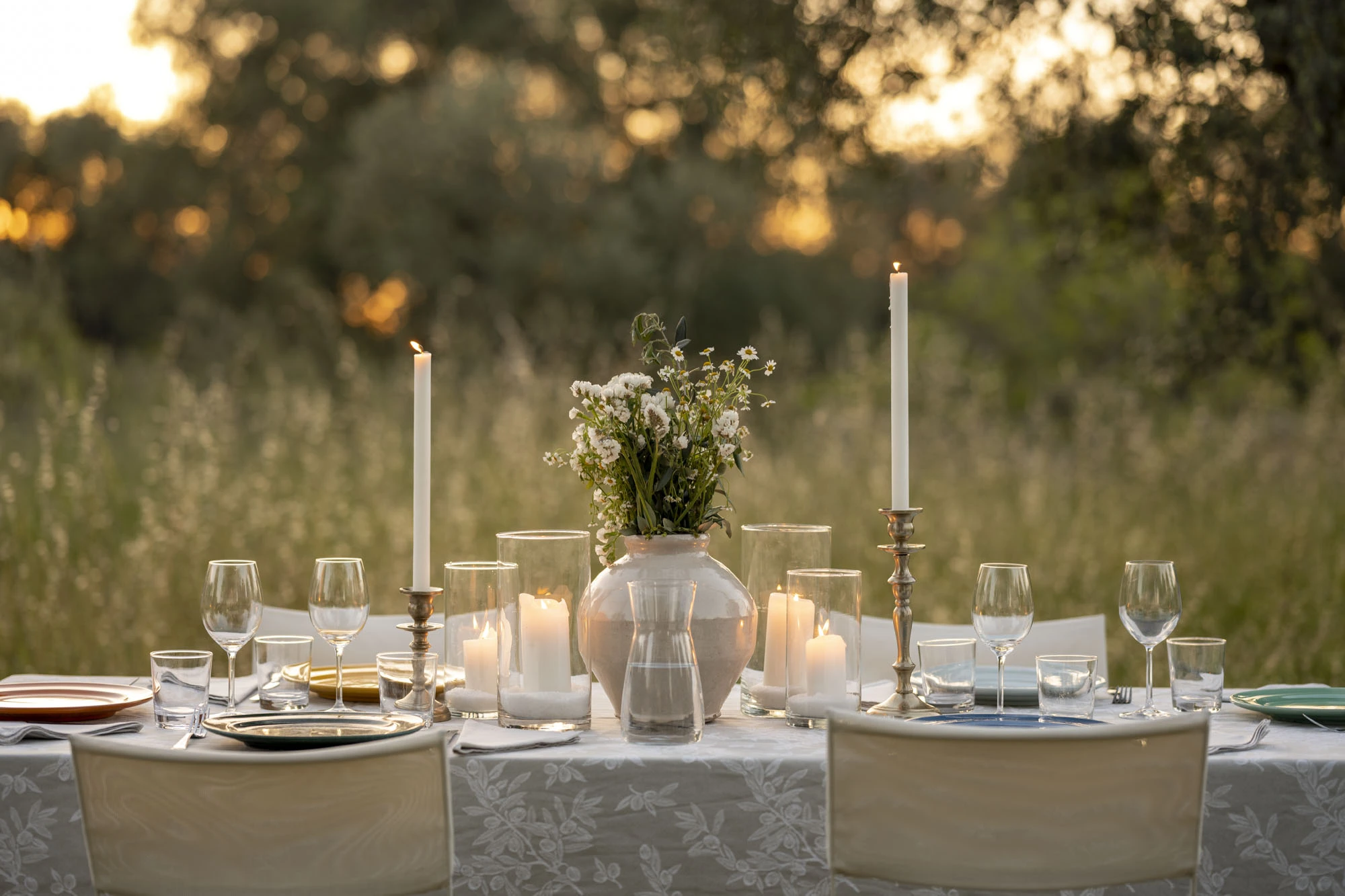 Elegantly set outdoor dinner table with white taper candles, hurricane lanterns, wildflower bouquet in ceramic vase, crystal glasses and coloured plates at golden hour in a Puglia olive grove