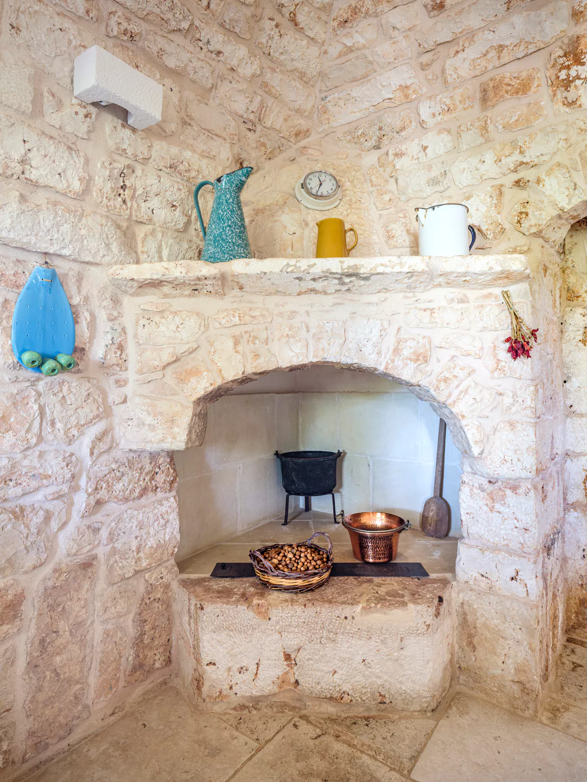Bright open-plan living and dining room with exposed wood beam ceiling, built-in fireplace with TV unit, large terracotta amphora, wicker baskets and floor-to-ceiling sliding glass doors opening to outdoor dining terrace at a Puglia villa