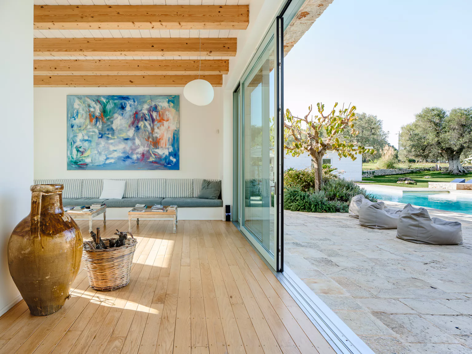 Bright contemporary living room with wood beam ceiling, large abstract painting, grey built-in sofa, terracotta amphora and floor-to-ceiling glass doors opening to pool terrace with olive trees at a Puglia villa