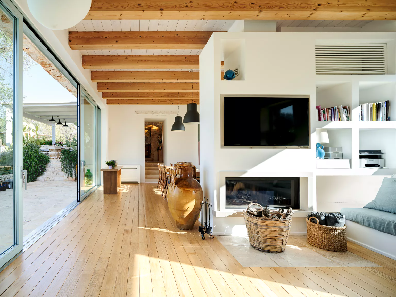 Bright open-plan living and dining room with exposed wood beam ceiling, built-in fireplace with TV unit, large terracotta amphora, wicker baskets and floor-to-ceiling sliding glass doors opening to outdoor dining terrace at a Puglia villa