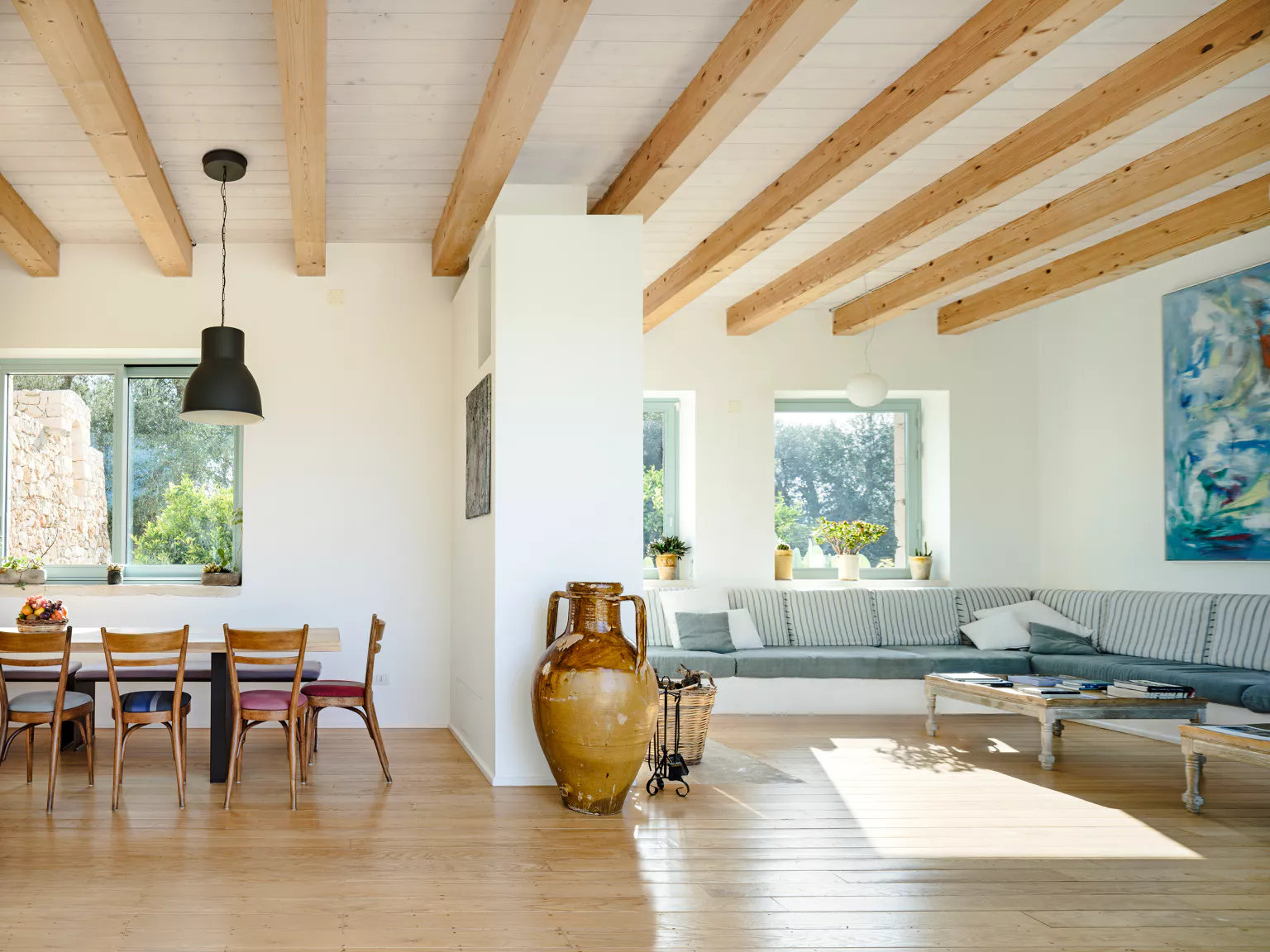 Wide open-plan living and dining room with white walls, exposed wood beam ceiling, grey built-in corner sofa with striped cushions, large amber amphora, abstract painting and garden views at a luxury Puglia villa