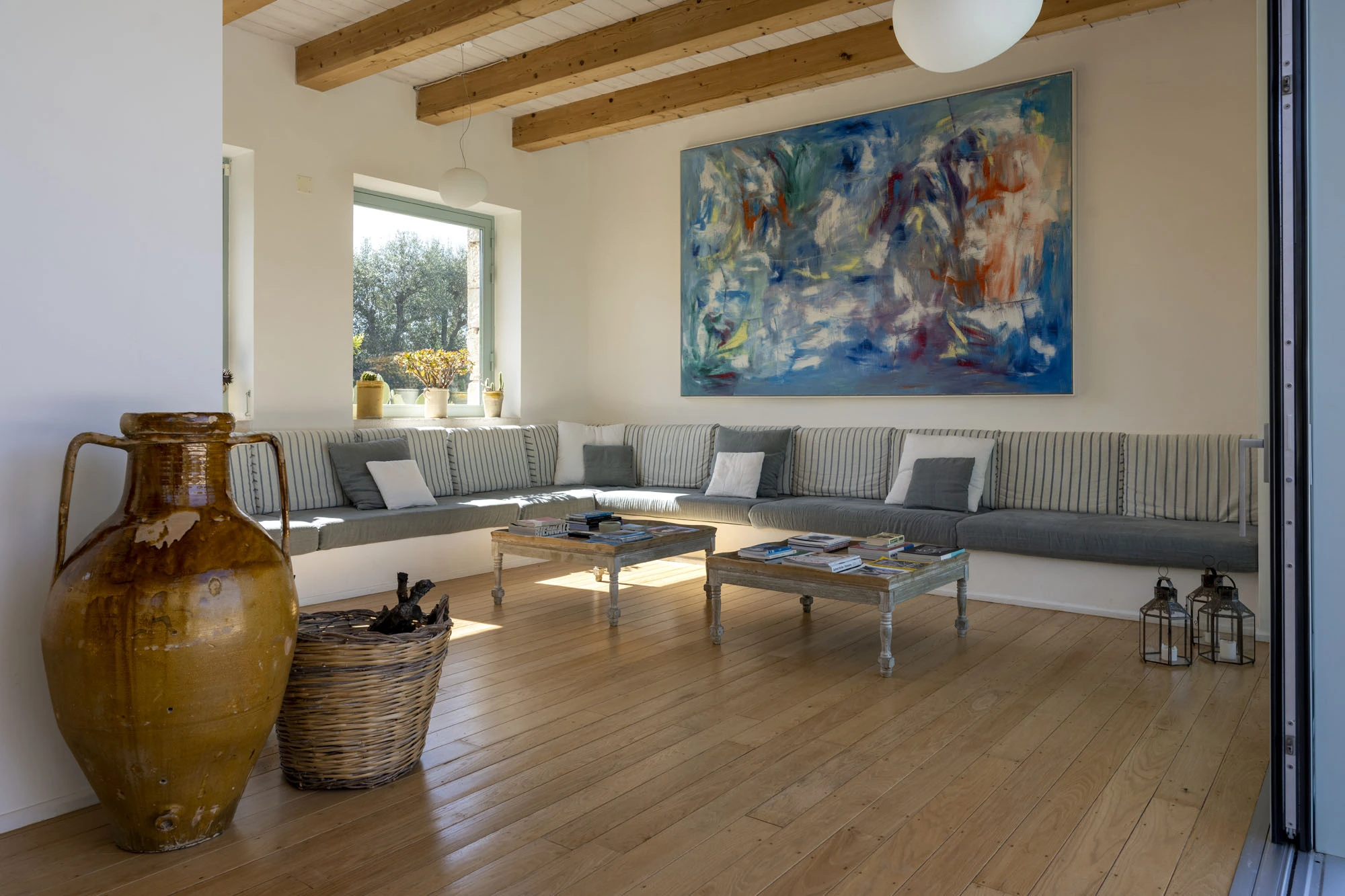 Generous living room with exposed wood beam ceiling, large grey built-in corner sofa with striped cushions, oversized terracotta amphora, bleached wood coffee tables and large abstract blue oil painting in a luxury Puglia villa