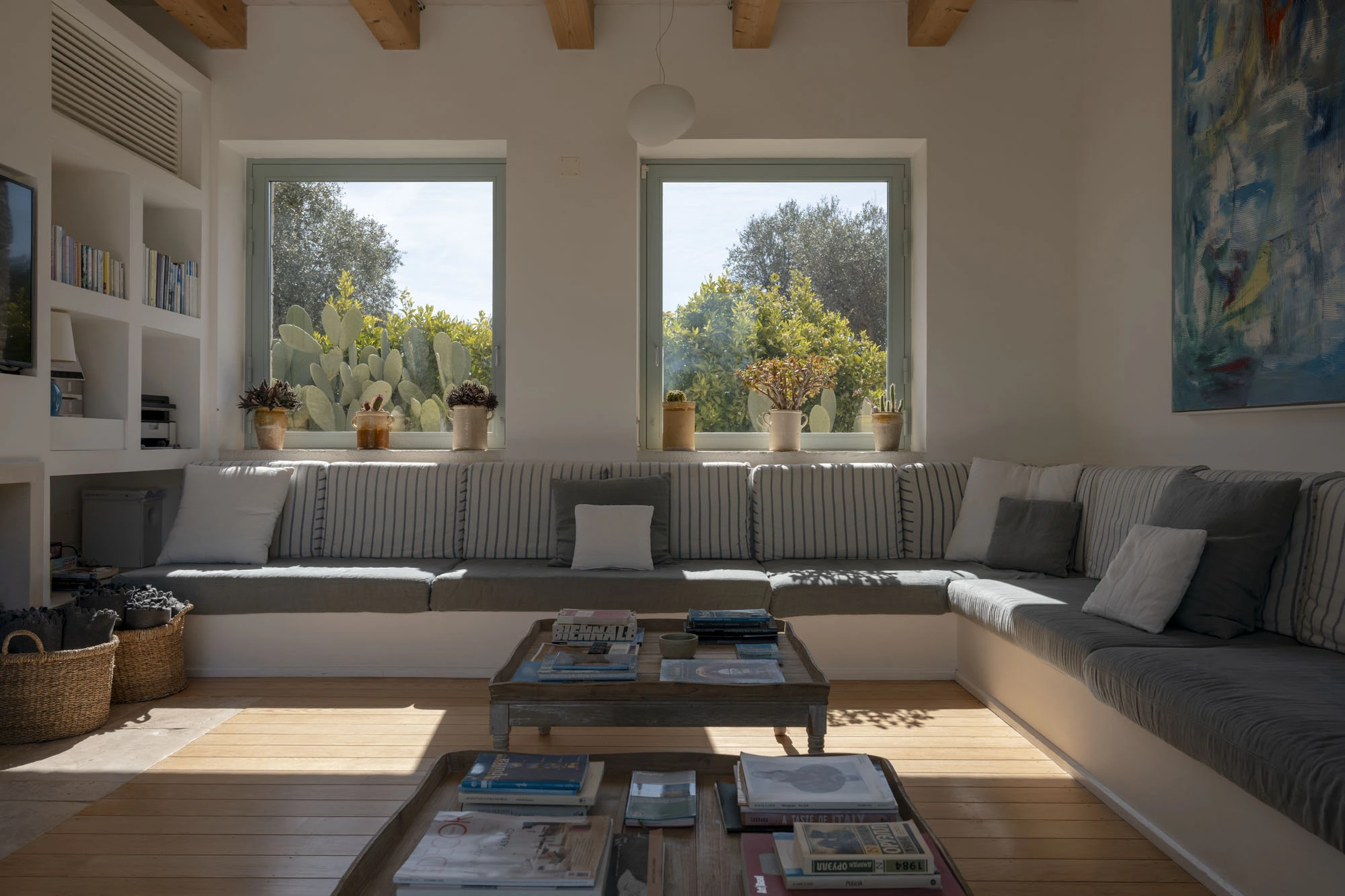 Bright living room with long built-in grey linen corner sofa, striped cushions, bleached wood coffee tables stacked with books, and twin windows overlooking prickly pear cacti and olive grove at a luxury Puglia villa