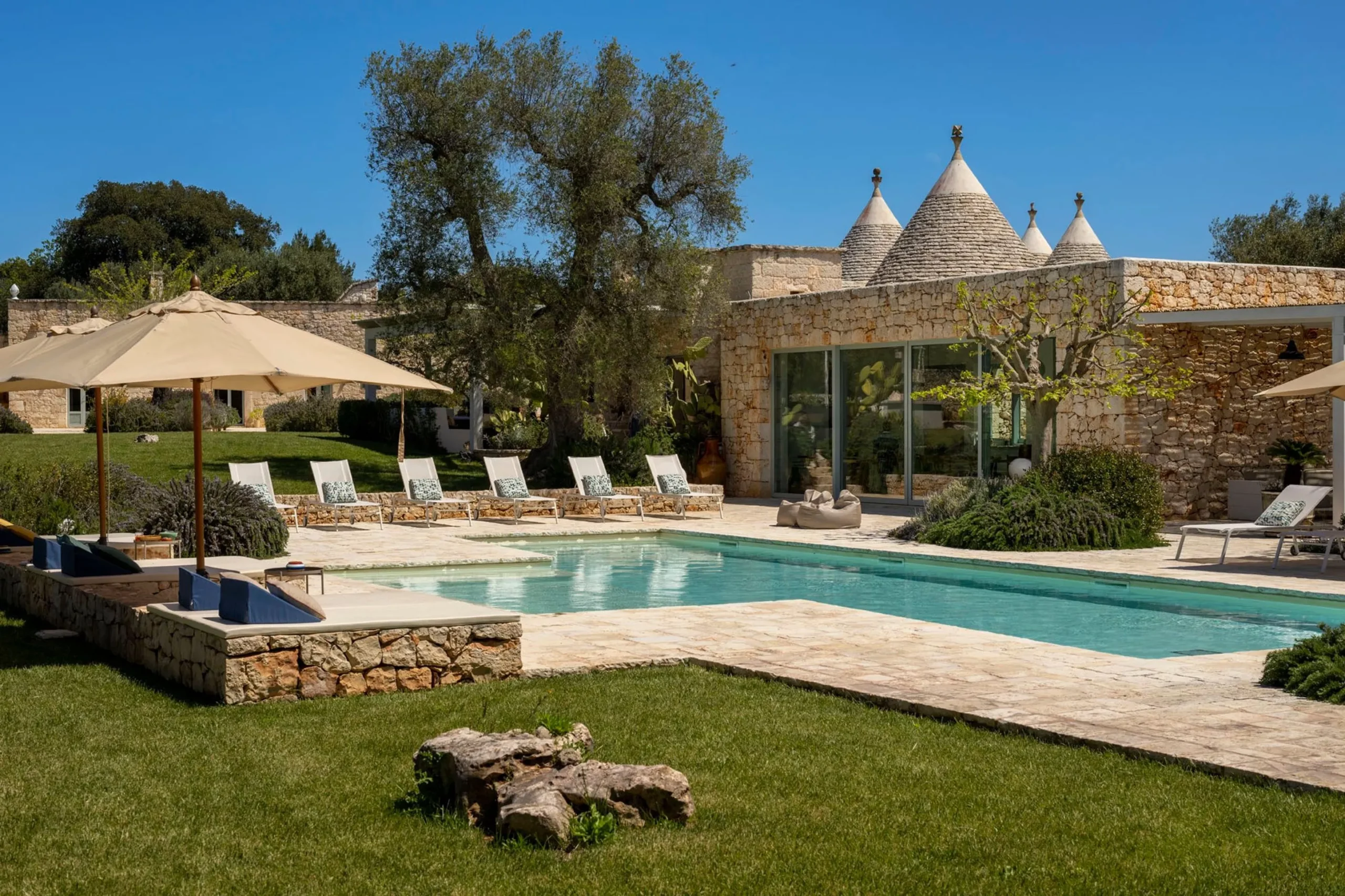Full view of a luxury Puglia trulli villa with large rectangular pool, white sun loungers with patterned cushions, stone terrace, bean bag chairs, lavender garden and ancient olive trees under a blue sky